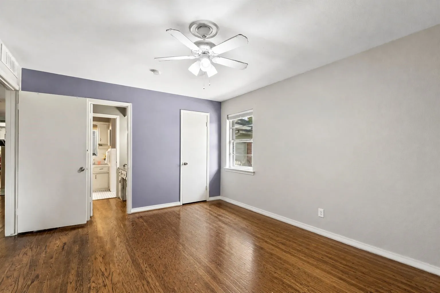 Spacious primary bedroom features wood flooring, ceiling fan with light fixture, a window with exterior views, and a closet with a solid door. Furniture removed by AI.