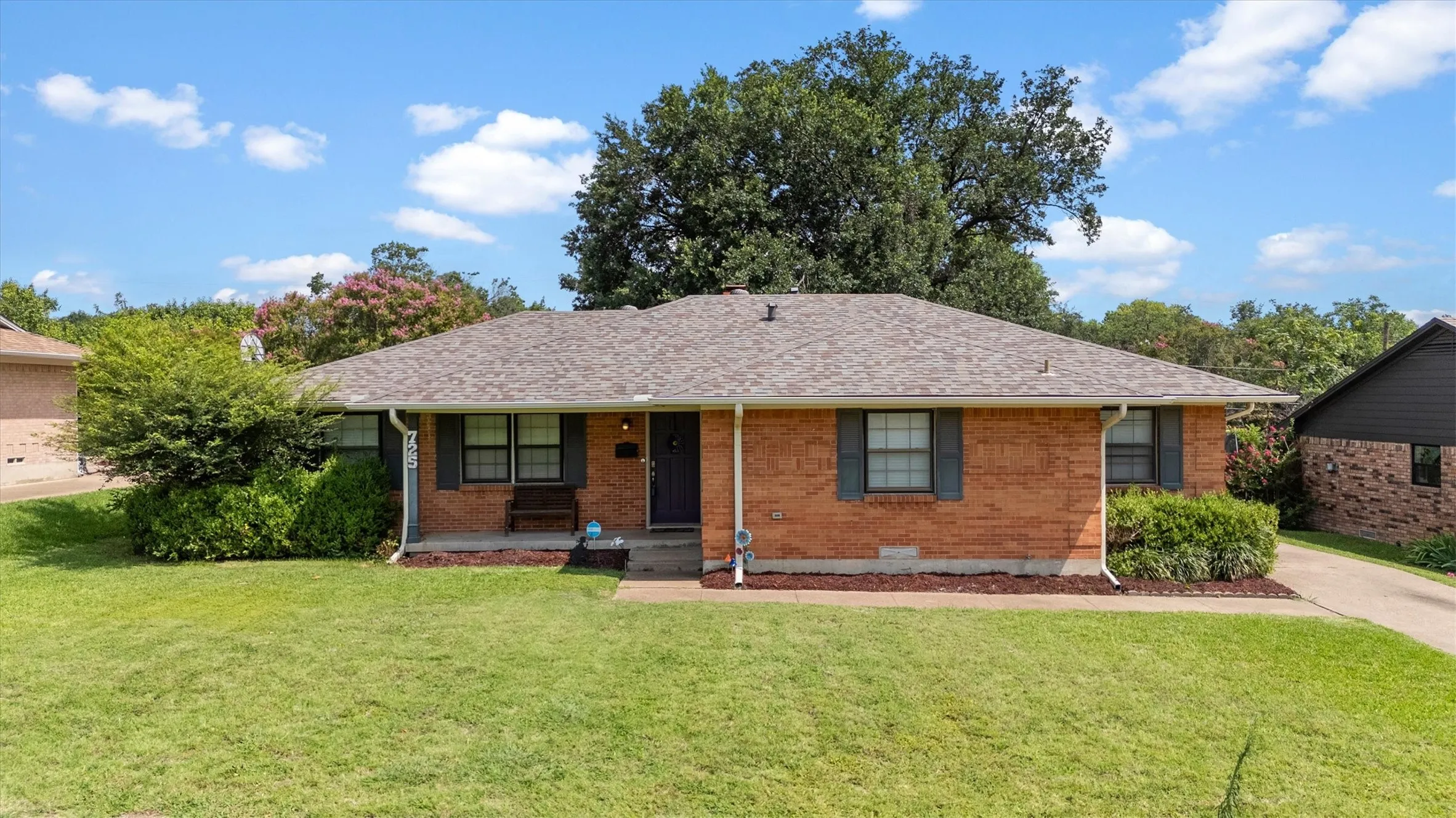 Brick home featuring a front porch, gabled roof, front lawn, and mature landscaping