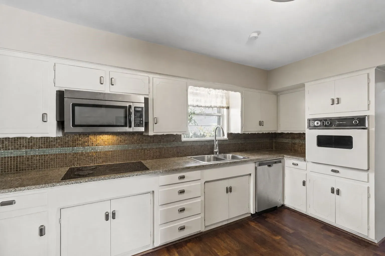 Kitchen featuring granite counters, mosaic tile backsplash, white cabinetry, a built-in stainless steel microwave, a built-in wall oven, a stainless steel dishwasher, stainless steel double basin sink and wood-finish flooring. Kitchen featuring rich wood-finish flooring, granite counters, mosaic tile backsplash, white cabinetry, integrated stainless steel dishwasher, and a stainless steel sink with a contemporary faucet. Wall decorations removed by AI.