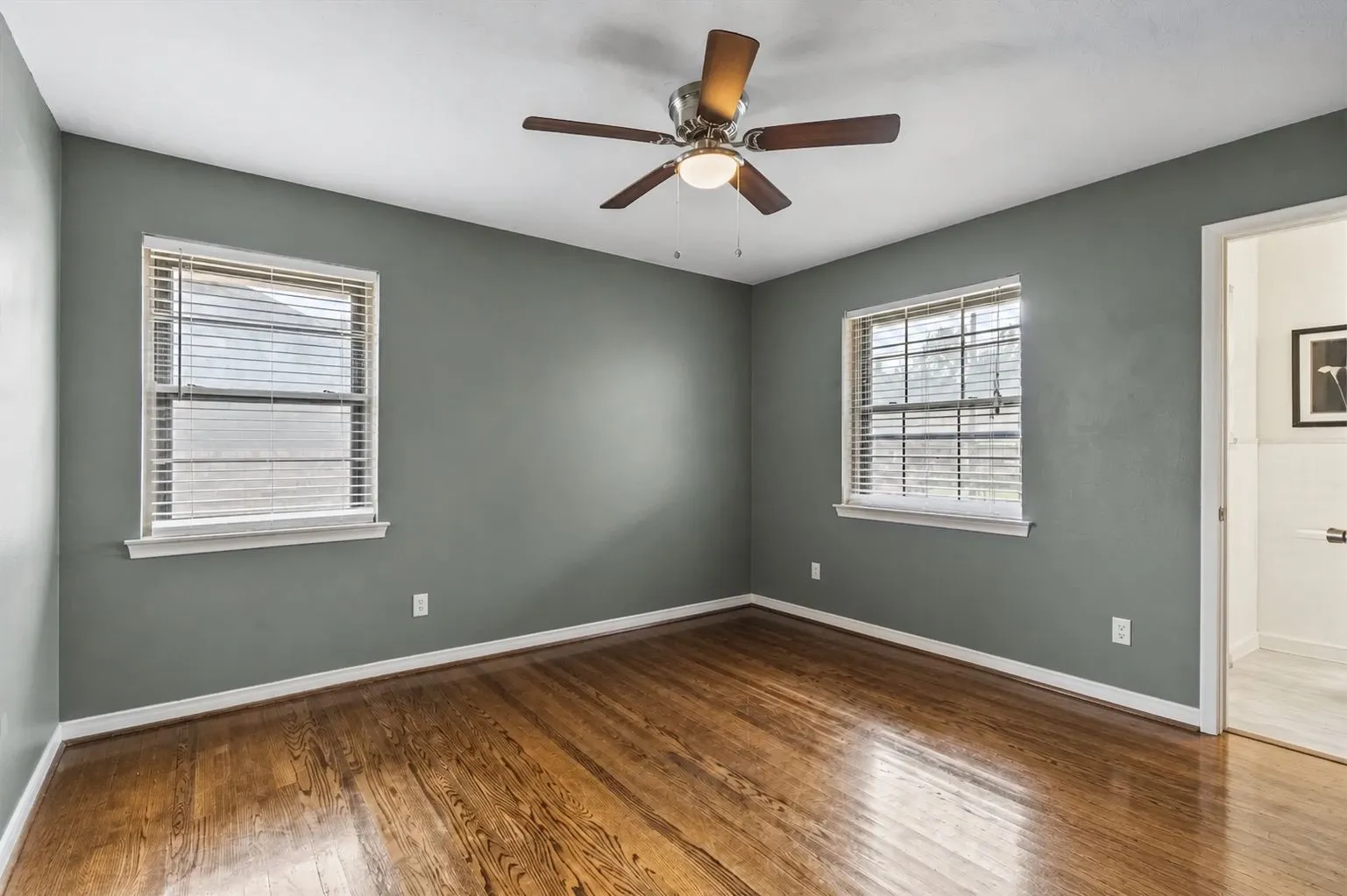 Bedroom featuring wood-finish flooring, 2 windows with horizontal blinds, and a ceiling fan with integrated lighting. Furniture removed by AI.