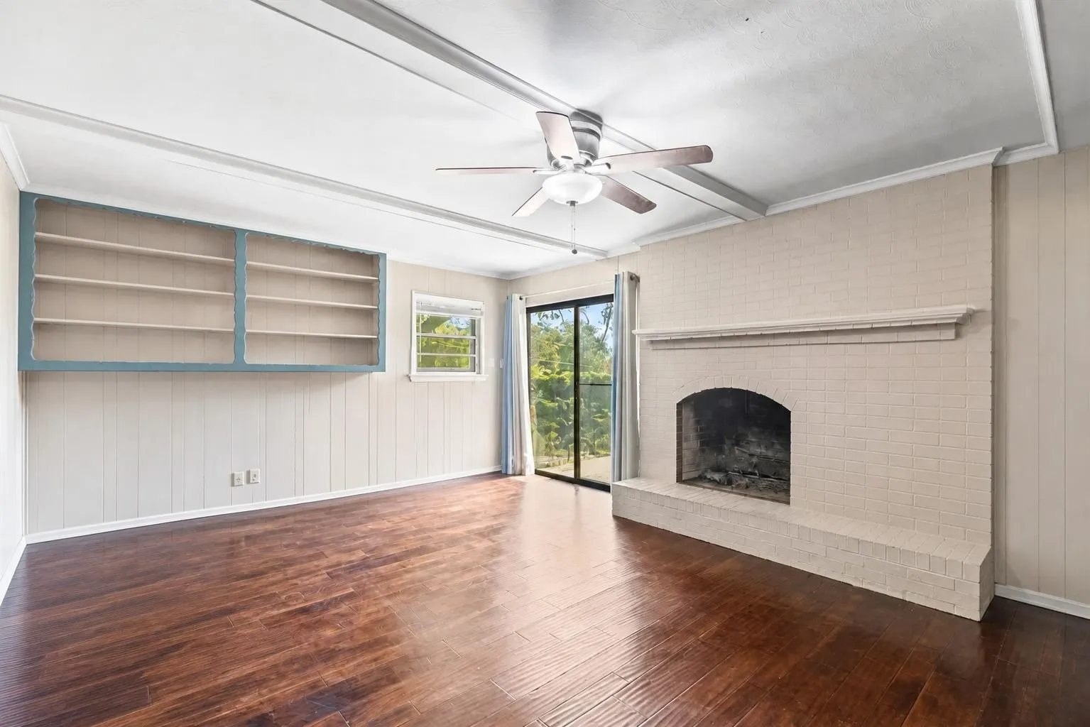 Living room featuring a brick fireplace with a mantle, dark wood-finish flooring, a ceiling fan with light fixture, white paneled walls, built in shelves and a sliding glass door to an exterior patio. Furniture removed with AI.