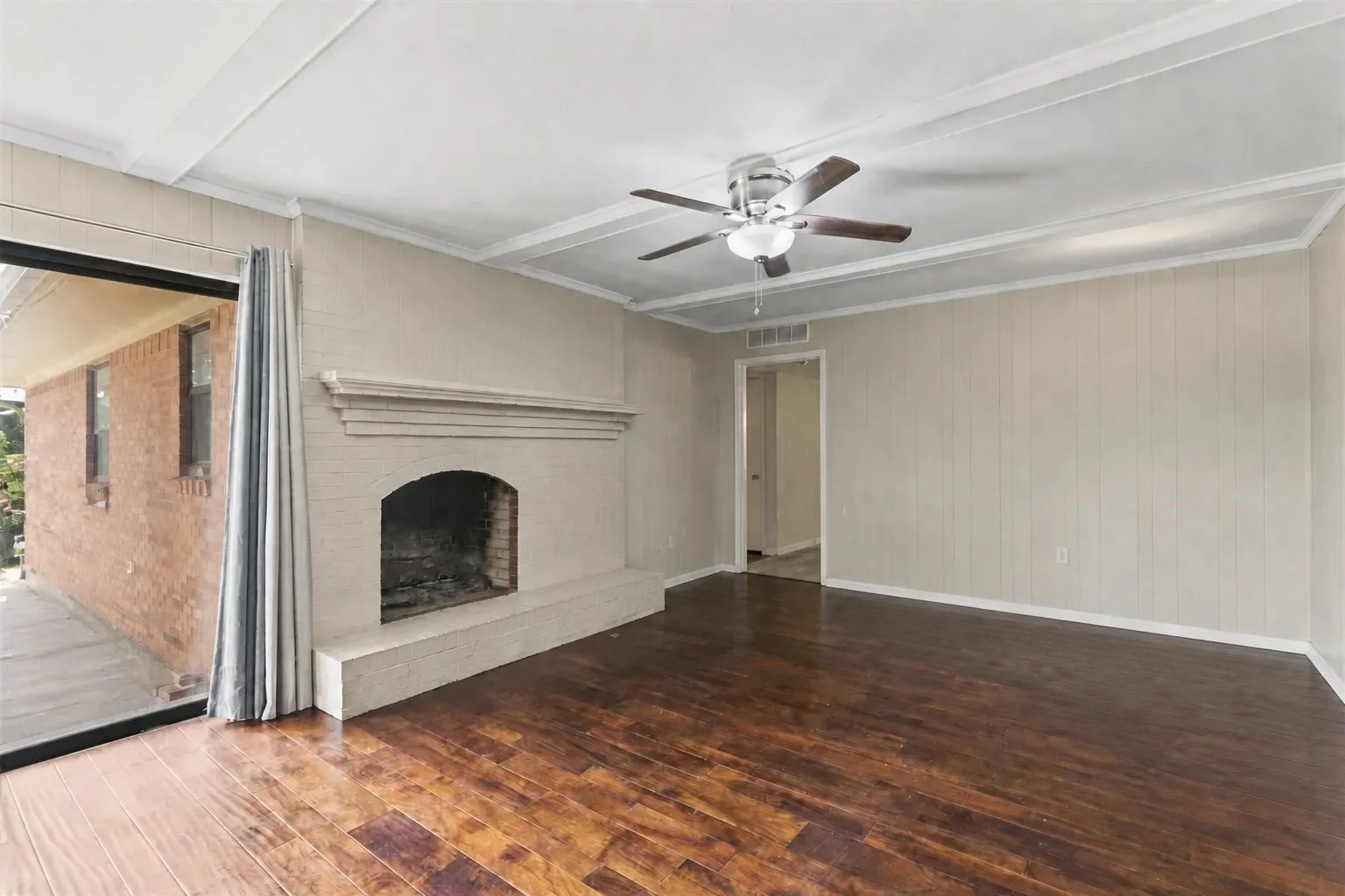 Living room featuring a brick fireplace with a mantle, dark wood-finish flooring, a ceiling fan with light fixture, white paneled walls, and a sliding glass door to an exterior patio. Furniture removed with AI.