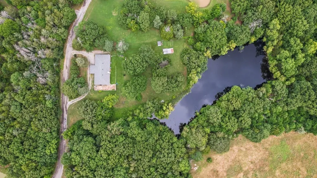 Expansive lot featuring a private pond, mature tree coverage, a main residence with a gray roof, and a gravel drive
