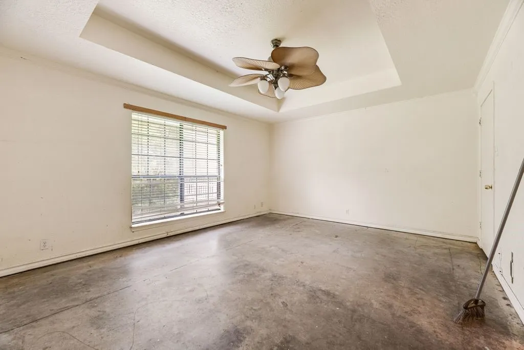 Tray ceiling with a ceiling fan light fixture, a large window with blinds, and concrete flooring