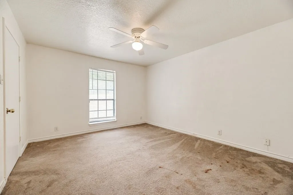 Spacious room featuring neutral carpeting, white walls, a single window with blinds, a white ceiling fan with integrated lighting, and a white interior door