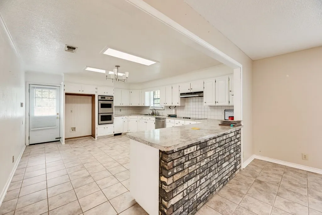 Spacious kitchen area featuring tile flooring, white cabinetry, integrated double wall ovens, a tile backsplash, and a breakfast bar with a brick facade base