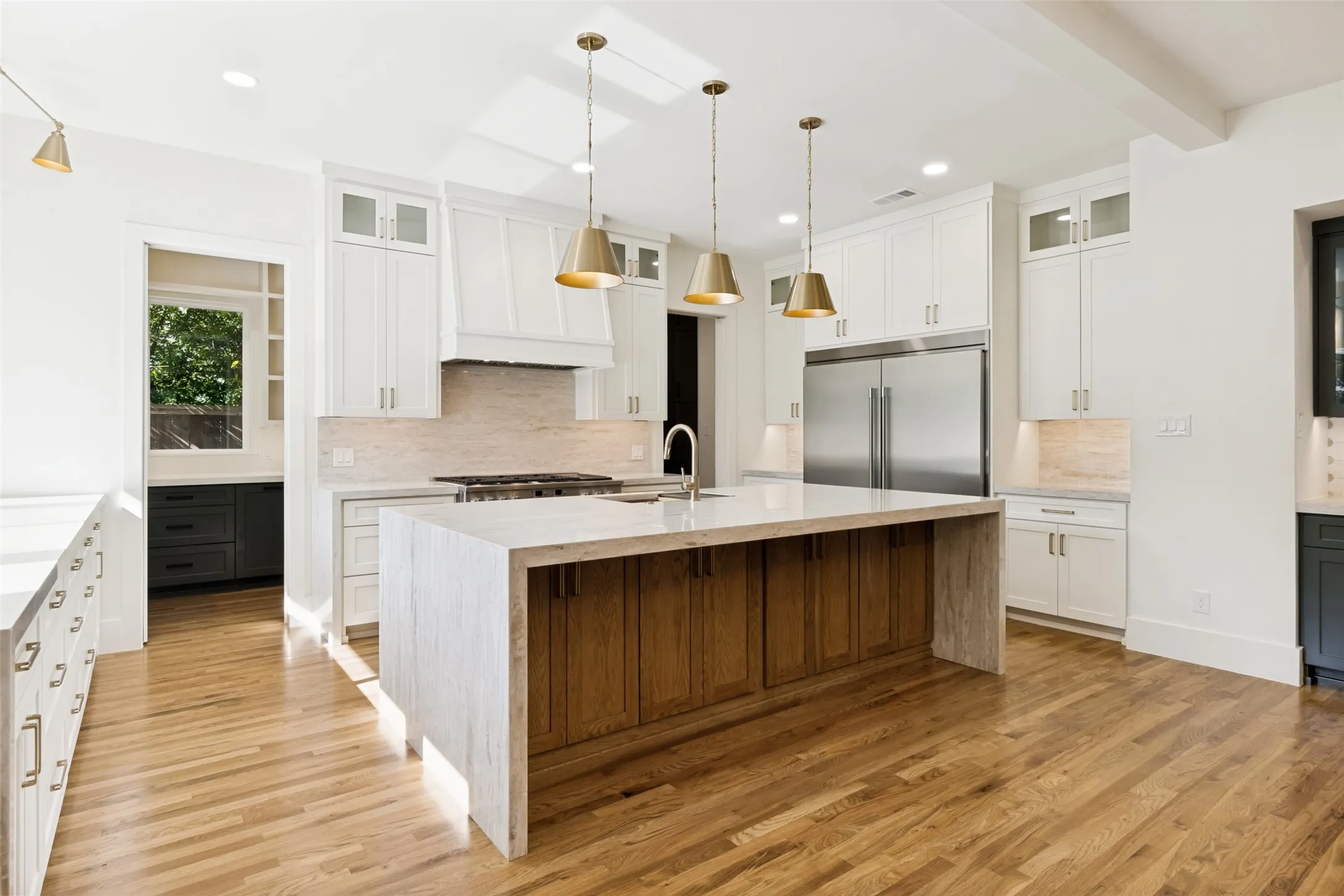 Spacious kitchen featuring a large waterfall island with wood-finish base, stainless steel appliances, white cabinetry, brass pendant lighting, and wood-finish flooring