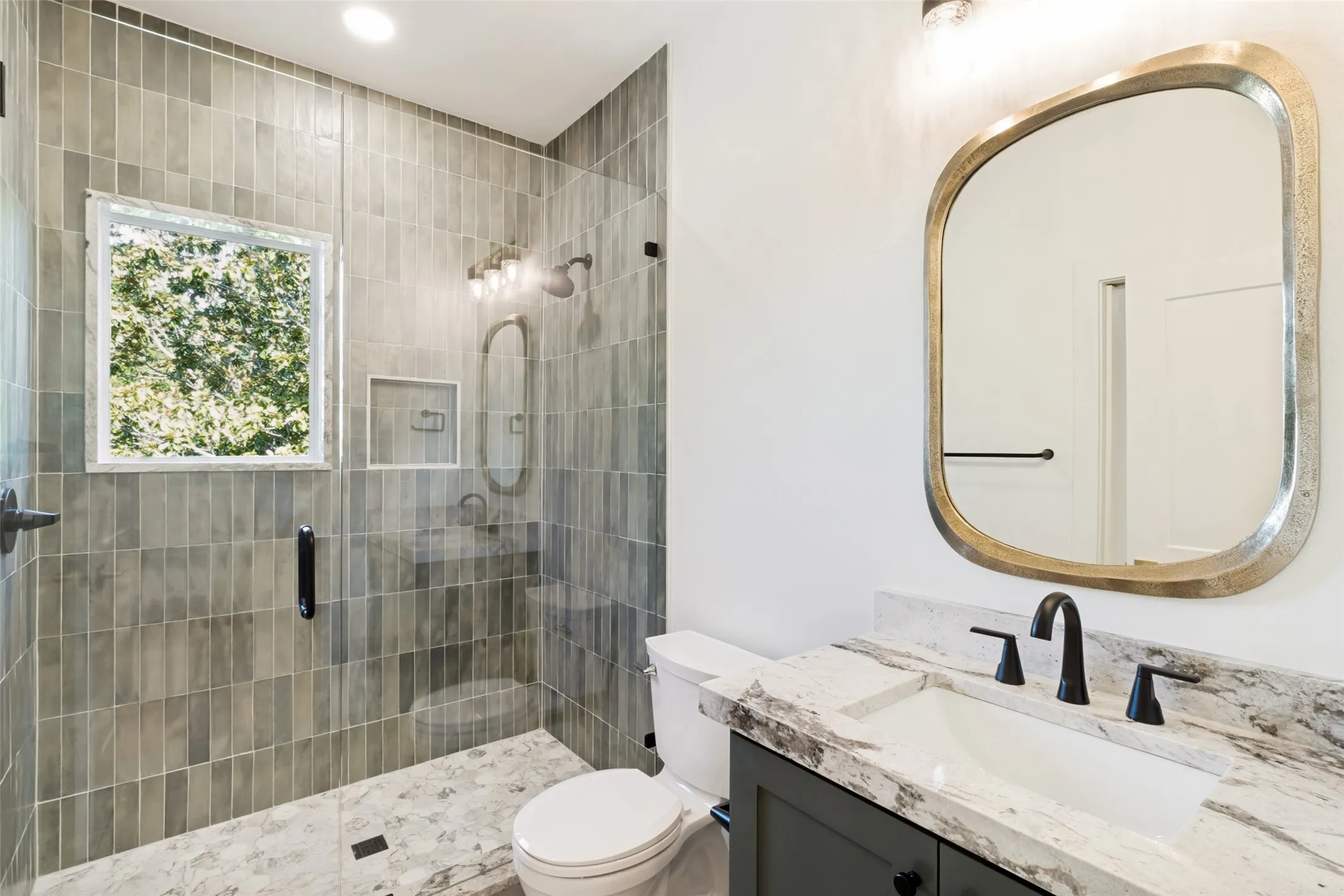 Bathroom featuring a walk-in shower with vertical tile and glass enclosure, a white vanity with stone countertop and integrated sink, a rectangular mirror, and matte black fixtures