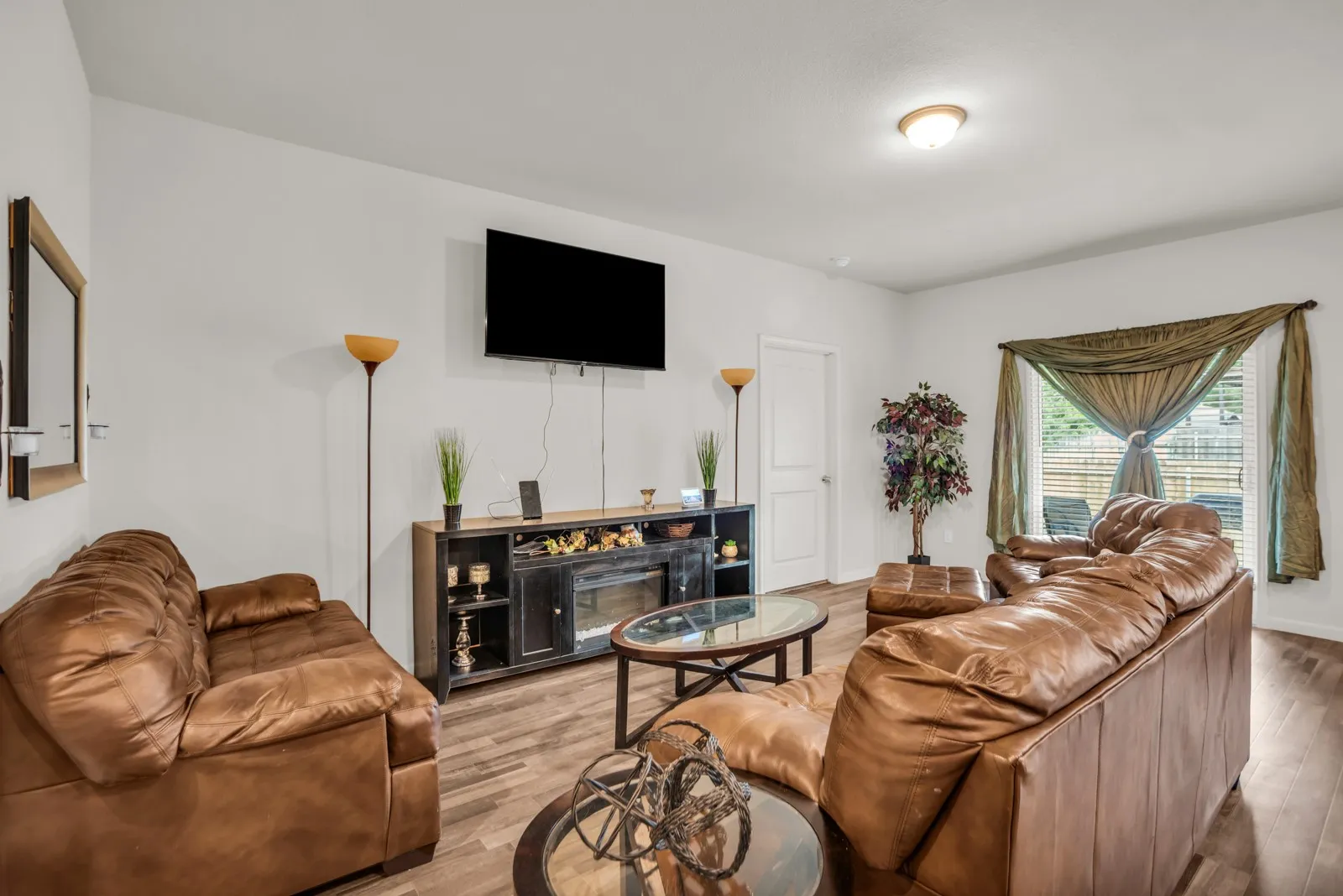Spacious living area featuring wood-finish flooring, white walls, a mounted television, a built-in media console with a fireplace insert, and a large window with blinds