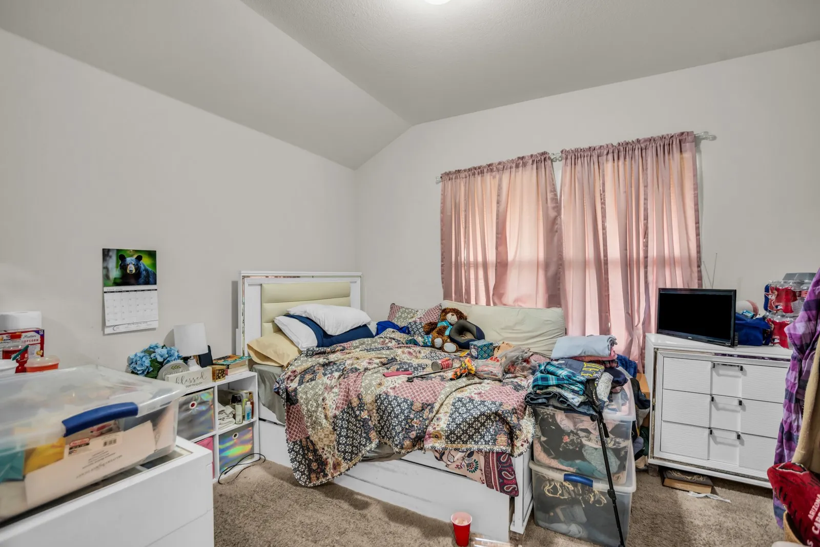 Carpeted room featuring a window with privacy curtains, a white bed frame with a padded headboard, and a vaulted ceiling