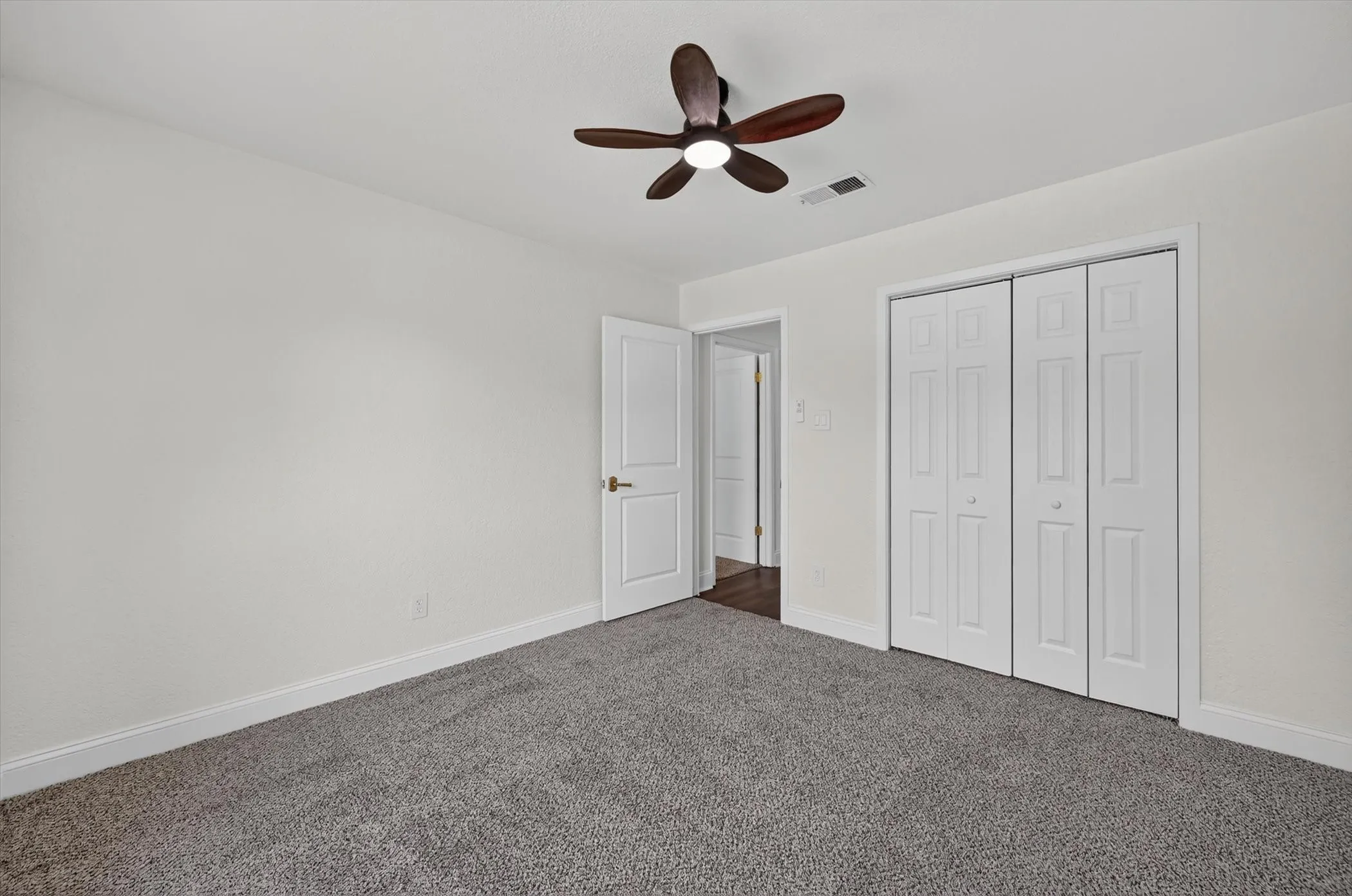 Interior room featuring neutral wall paint, gray carpeting, a dark wood-finish ceiling fan with integrated lighting, and white bi-fold closet doors
