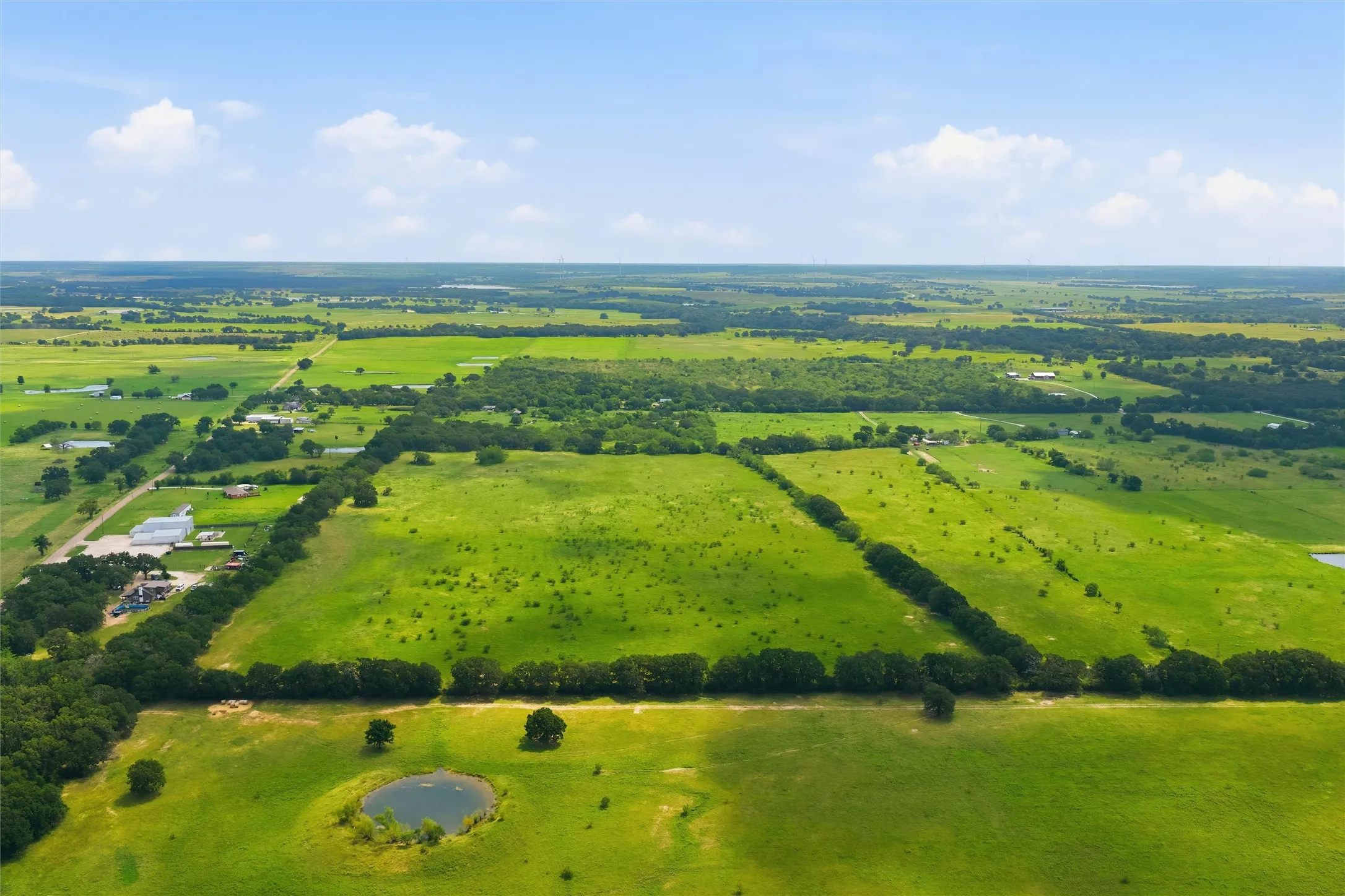 Expansive green acreage featuring a serene pond, mature tree lines, and open fields under a clear sky