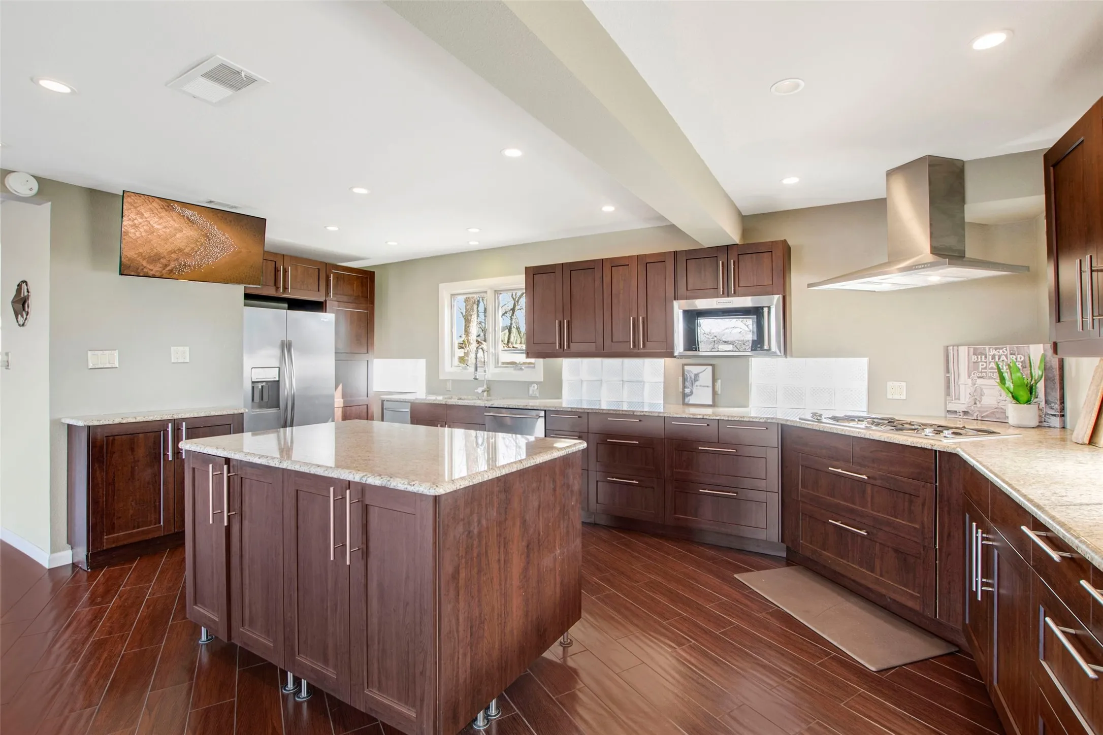 Kitchen featuring a central island with a light-toned countertop, rich wood-finish flooring, dark wood cabinetry, and stainless steel appliances including a refrigerator, microwave, and range hood