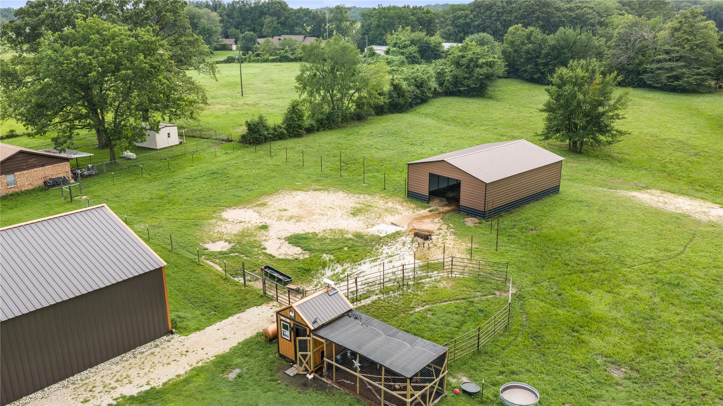 Expansive green property featuring multiple outbuildings, including a large brown metal barn with a matching roof, a smaller brown metal shed with a grey roof, and a wooden chicken coop with an attached fenced enclosure