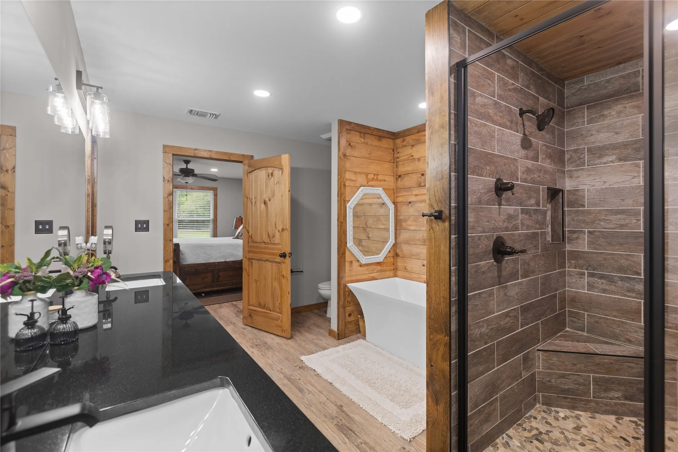 Spacious bathroom featuring a freestanding soaking tub, walk-in shower with tiled walls and pebble flooring, dual vanity with a dark countertop, and wood-finish flooring