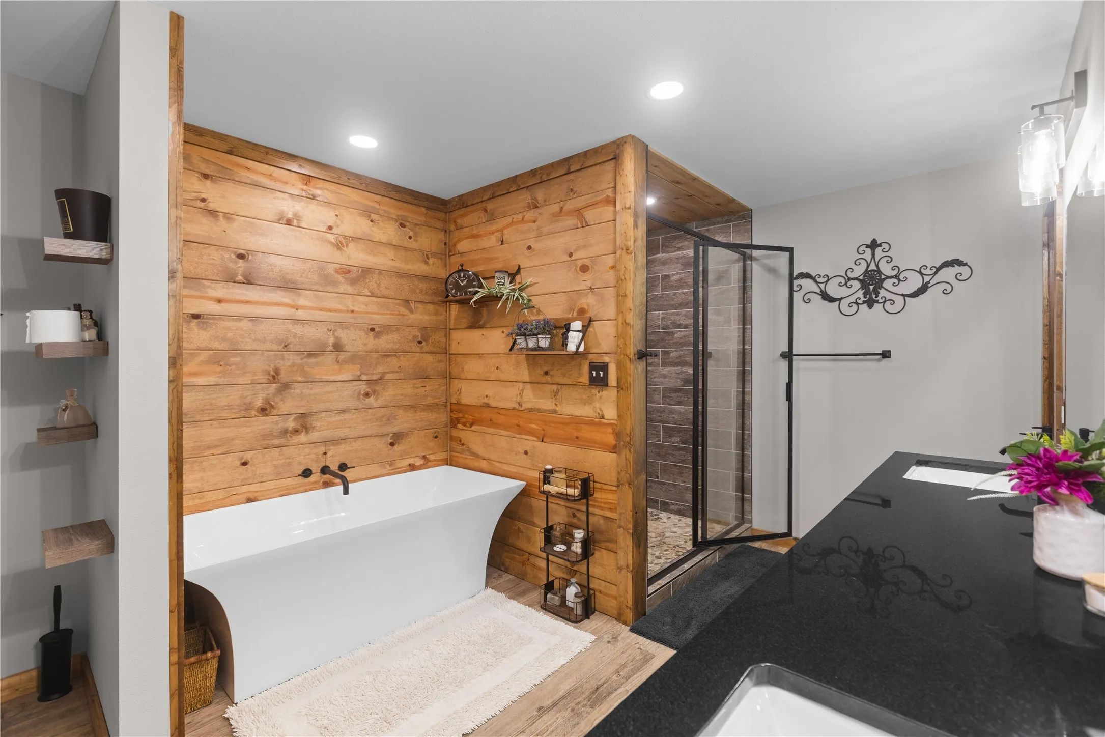 Bathroom featuring wood plank accent walls, a freestanding soaking tub with matte black fixtures, a walk-in shower with a glass door and stone-finish tile surround, and a dark stone-finish double vanity