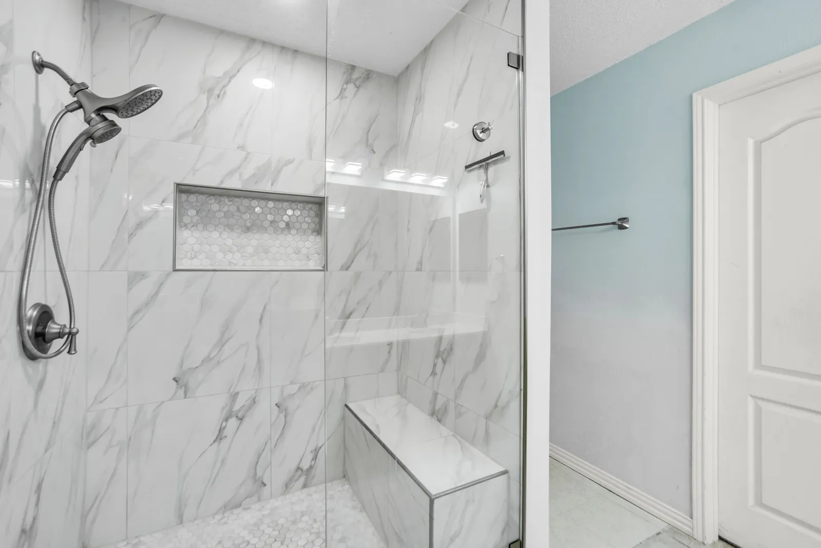 Modern shower enclosure featuring a glass door, marble-look tile walls, a built-in shower niche with hexagonal tile, a built-in bench, and a dual-head shower system