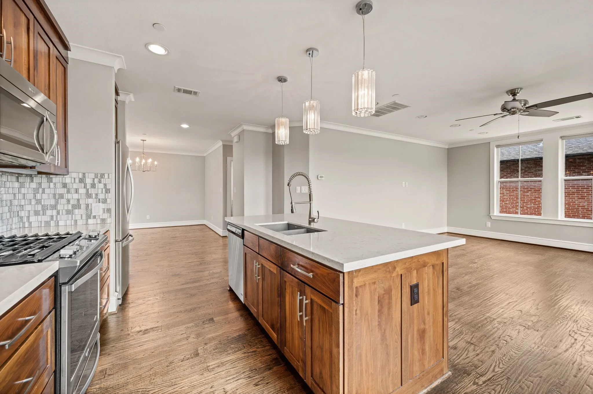 Open-concept kitchen featuring an island with a stainless steel sink and modern pendant lighting, stainless steel appliances, a mosaic tile backsplash, and wood-finish flooring extending into the adjacent living space