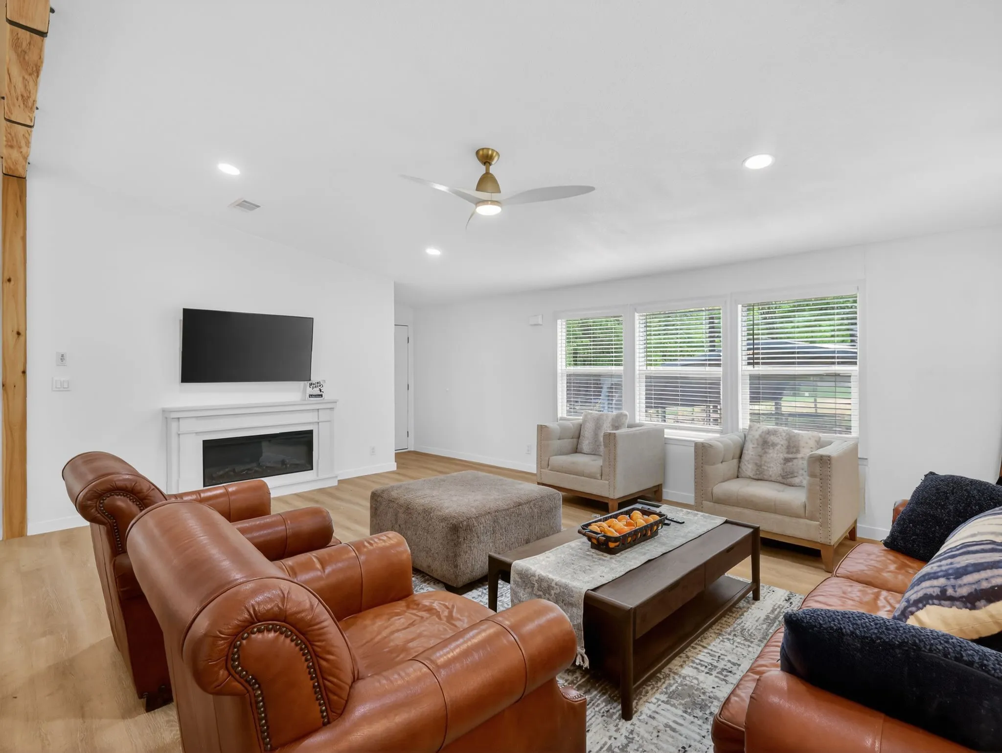 Spacious living area featuring light wood-finish flooring, a white mantel fireplace, and multiple windows with horizontal blinds