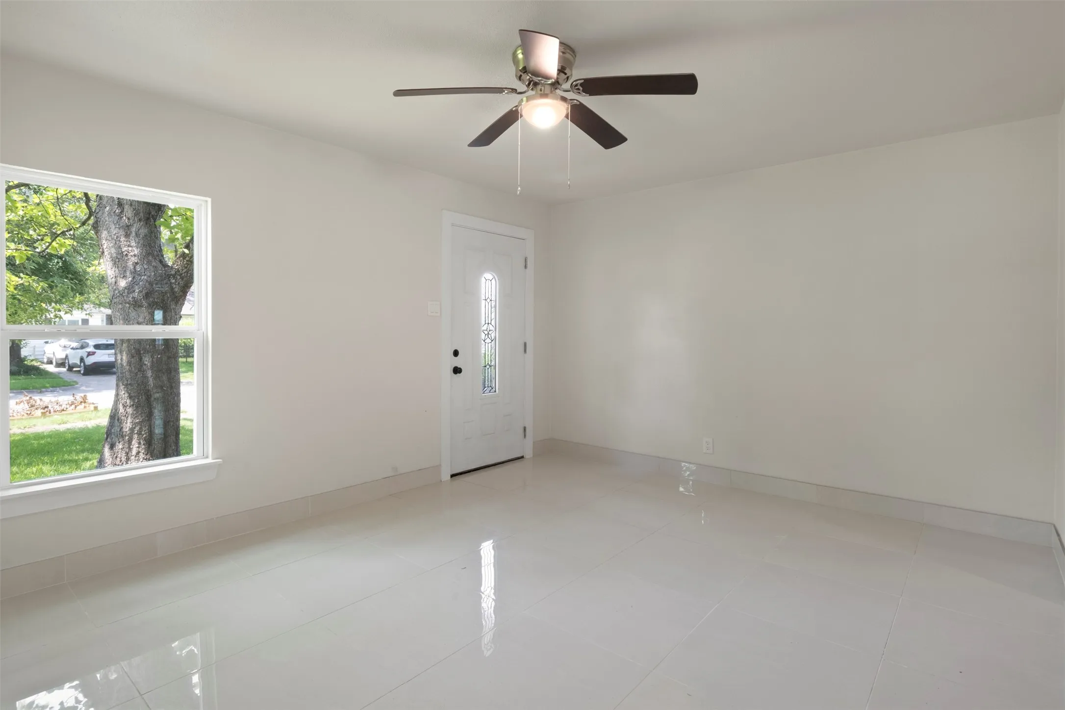 Spacious room featuring light-toned flooring, a ceiling fan with integrated lighting, a white entry door with decorative glass, and a large window providing outdoor illumination