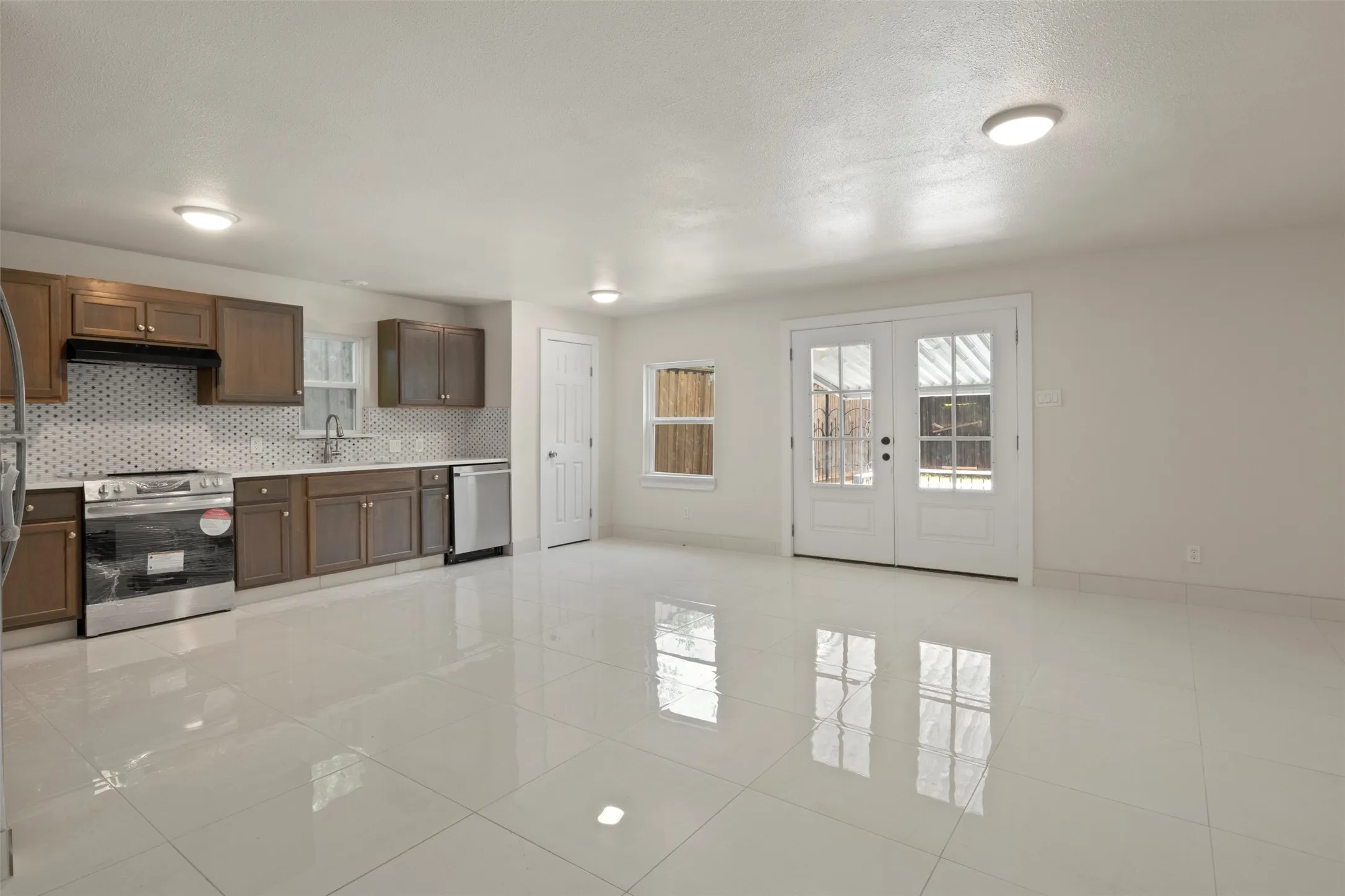Open-concept kitchen and living area featuring white tile flooring, brown cabinetry, stainless steel appliances, and a mosaic tile backsplash