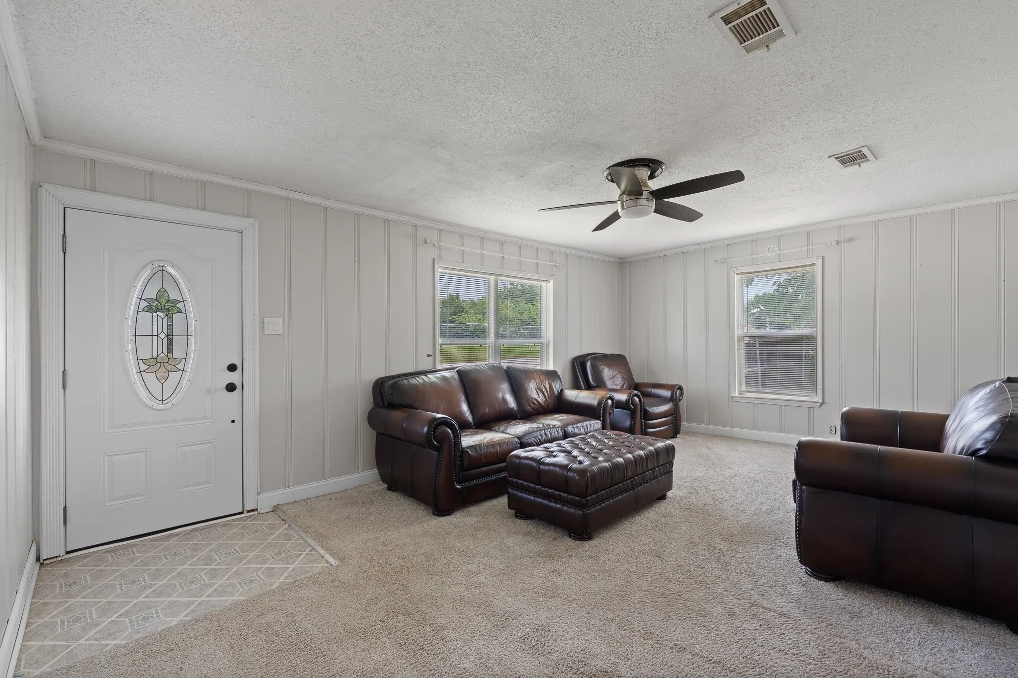 Living space featuring a white entry door with an oval decorative glass insert, board and batten wall paneling, two windows with exterior views, a ceiling fan, and carpet flooring