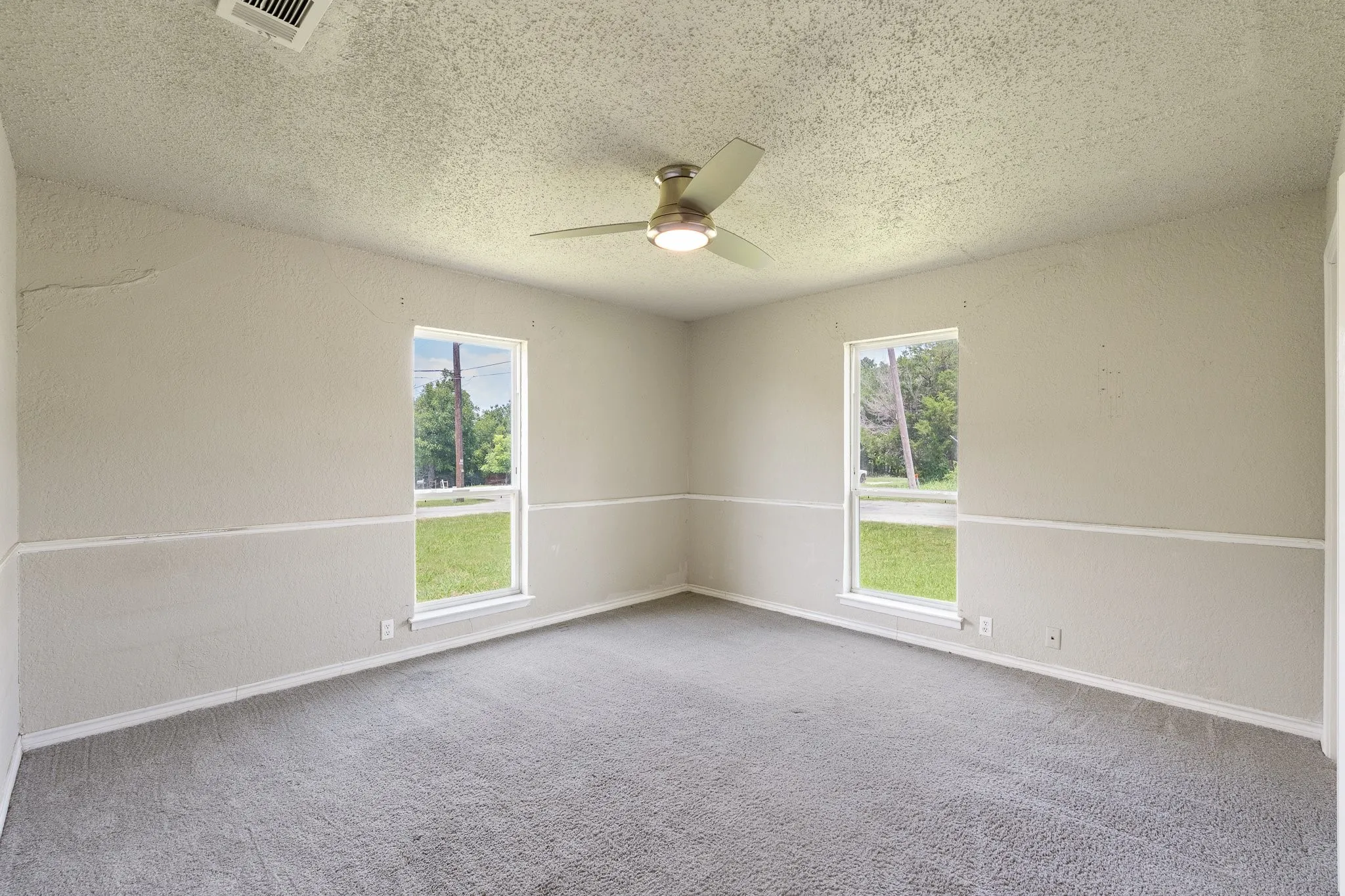 Room featuring two windows, neutral gray carpeting, a textured ceiling, a ceiling fan with integrated lighting, and painted walls with a chair rail detail
