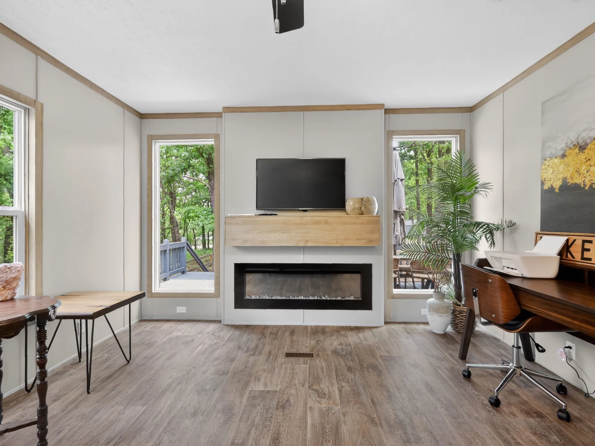 Contemporary living area featuring wood-finish flooring, an electric fireplace with a light wood mantle, two large windows, and neutral wall paneling