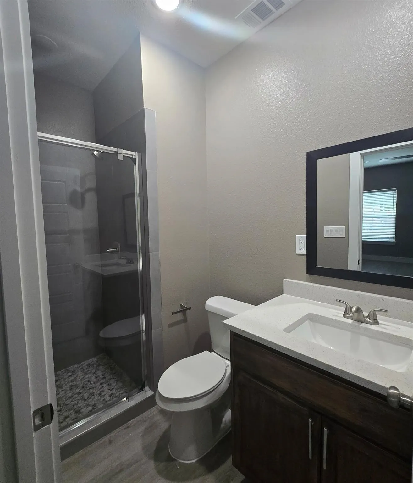 Contemporary bathroom featuring a glass-enclosed shower with pebble-style floor, a dark wood-finish vanity with a white countertop and integrated sink, brushed nickel fixtures, and wood-finish flooring
