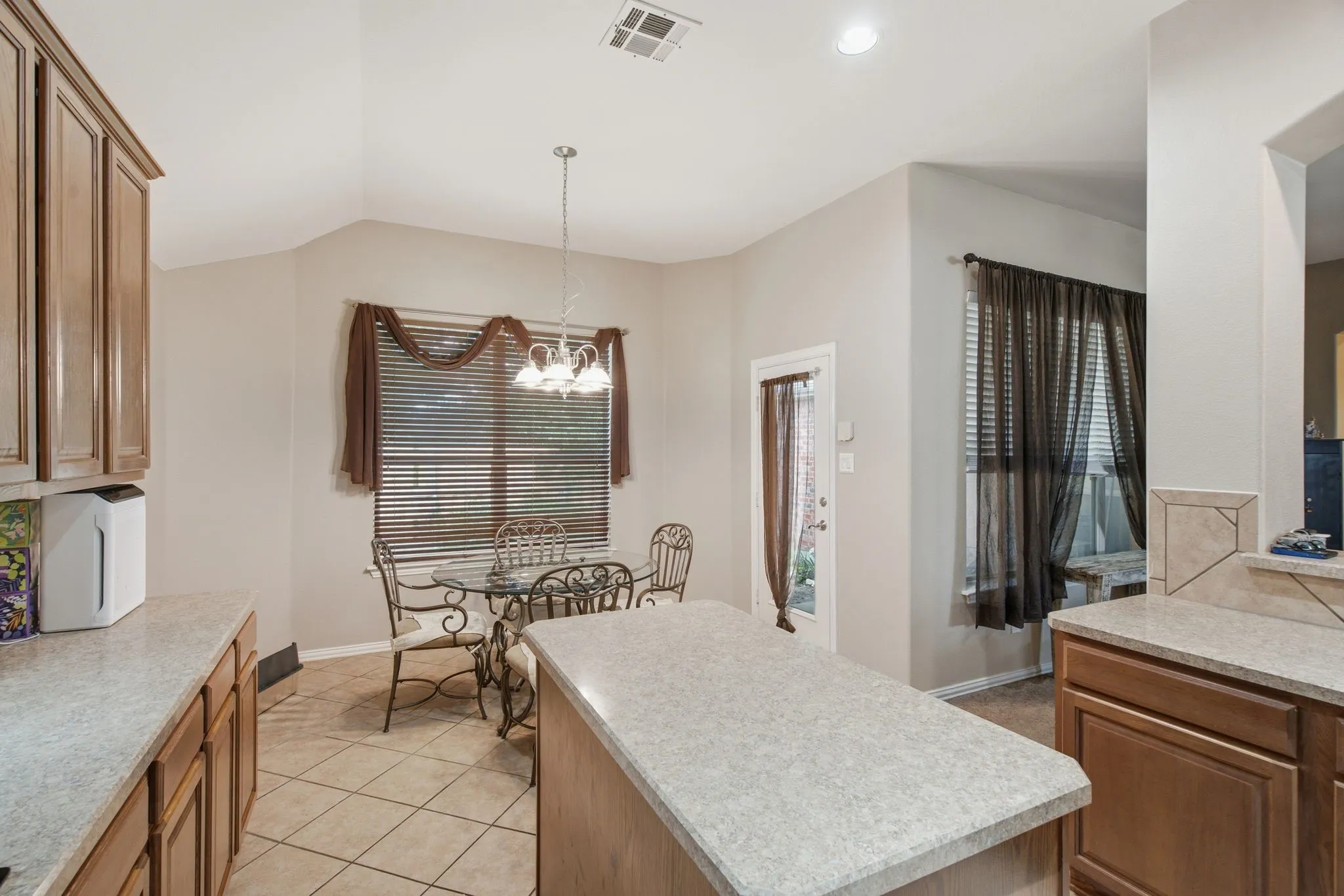 Kitchen featuring a central island with a speckled countertop, extensive wood-finish cabinetry, tile flooring, and a dining area with a window and a glass-paneled door