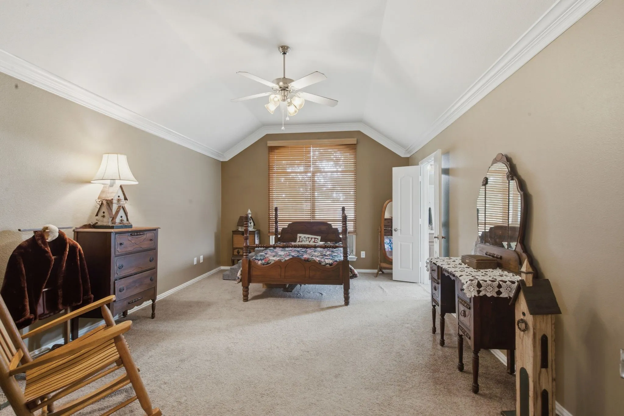 Carpeted master/primary bedroom featuring vaulted ceilings, crown molding, a ceiling fan with integrated lighting, and a window with blinds