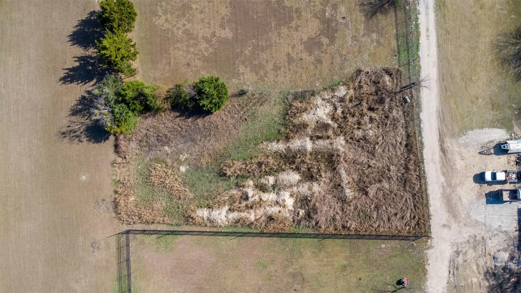 Expansive undeveloped land featuring a perimeter fence, scattered mature trees, and a gravel road bordering the property