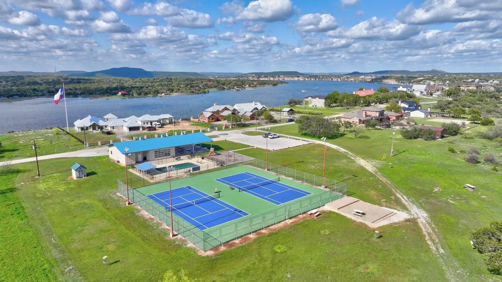 Aerial perspective showcasing dual tennis courts, a community building, and a swimming pool, all situated near a large body of water