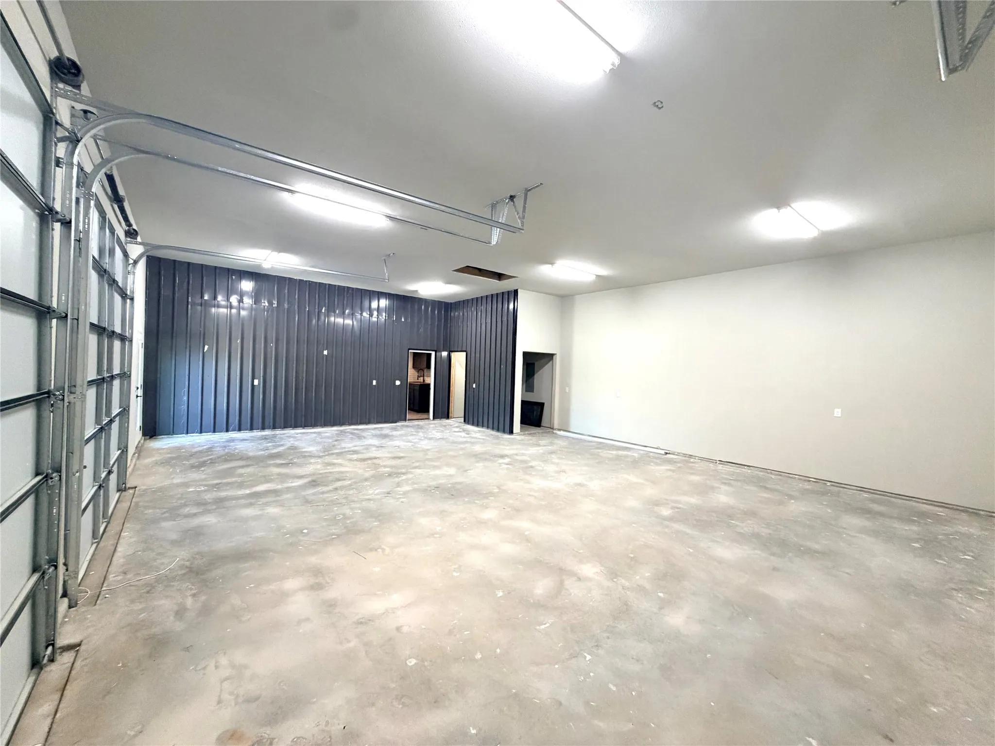 Spacious interior featuring a large roll-up door, concrete flooring, high ceilings, multiple fluorescent light fixtures, and a textured accent wall