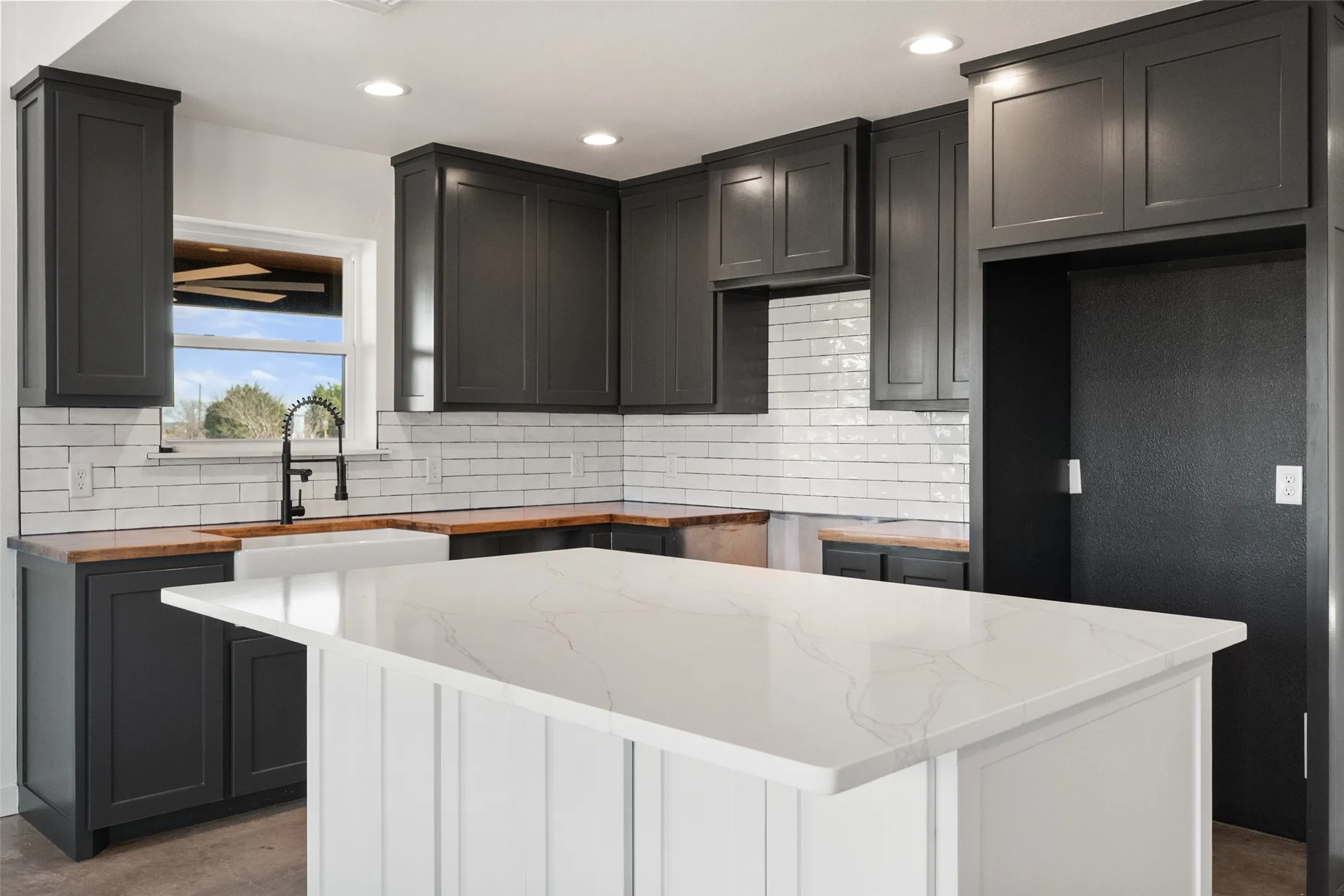 Modern kitchen featuring a central island with a white countertop, shaker-style dark cabinetry, butcher block countertops, a white subway tile backsplash, and a black gooseneck faucet with a farmhouse sink