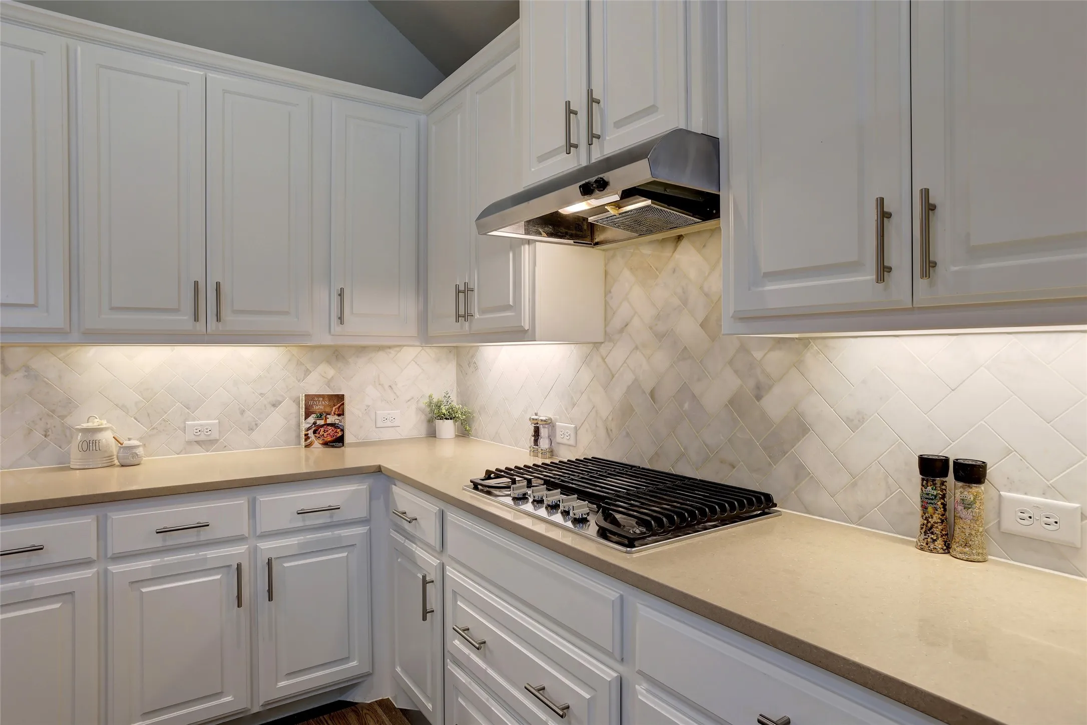 Kitchen featuring white cabinetry with brushed nickel hardware, stone-finish countertops, herringbone tile backsplash, a built-in gas cooktop, and under-cabinet lighting