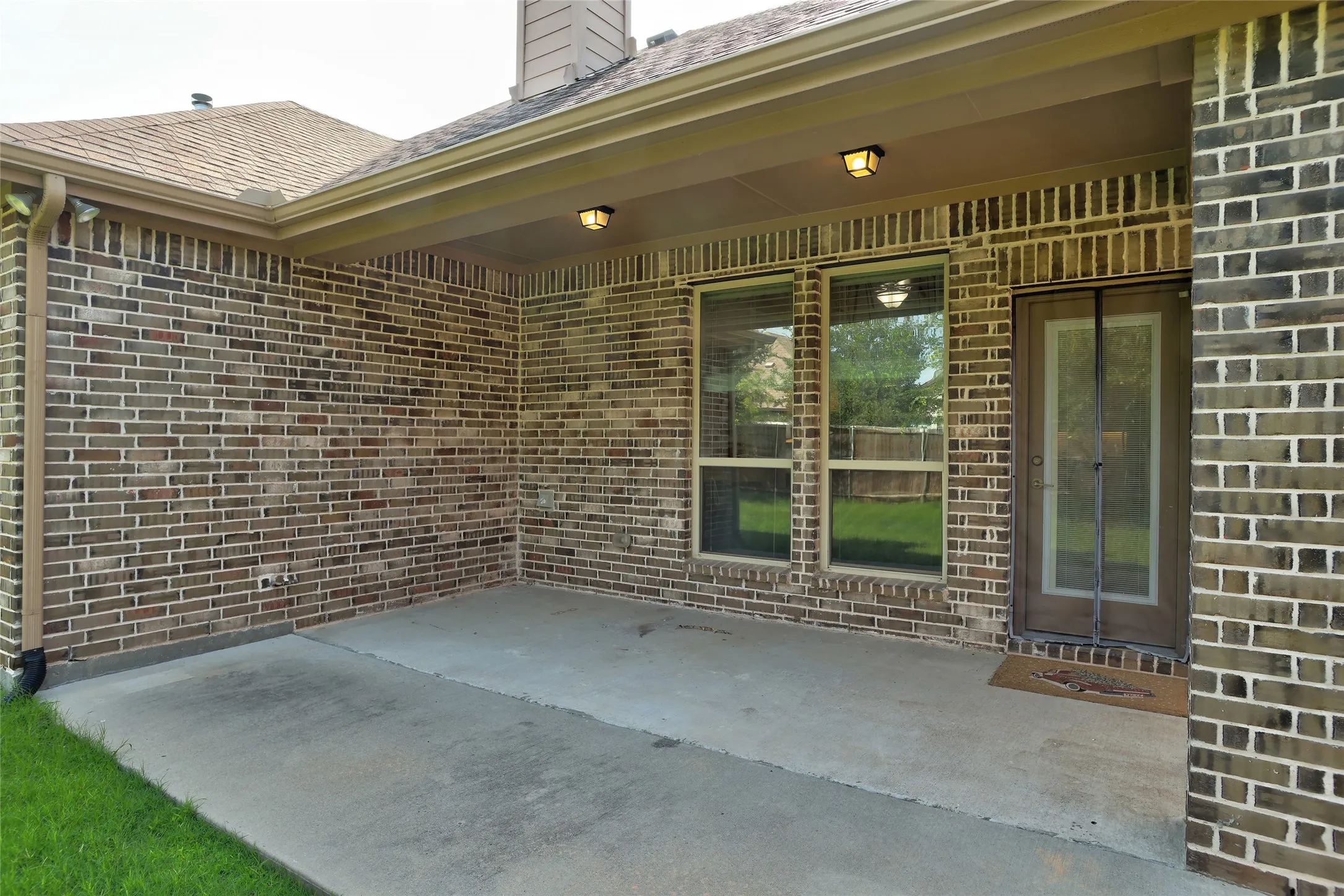 Covered concrete patio with brick exterior and dual windows, featuring a glass-paneled door