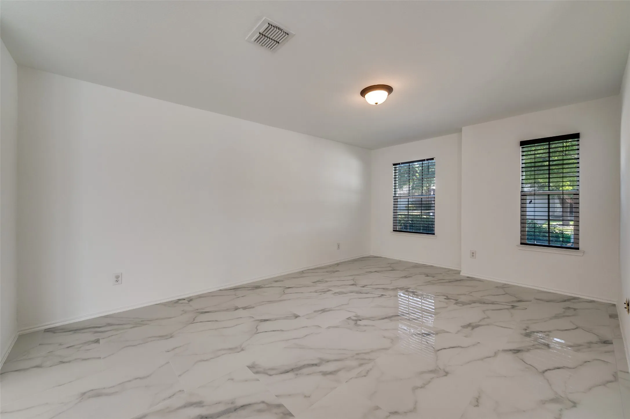 Spacious room featuring large format marble-look tile flooring, two windows with blinds, a ceiling-mounted light fixture, and white painted walls