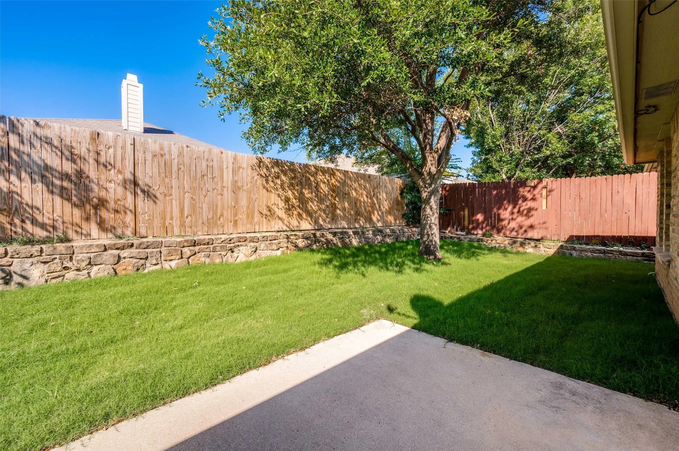 Private backyard featuring a concrete patio, a mature tree providing shade, and a stone retaining wall