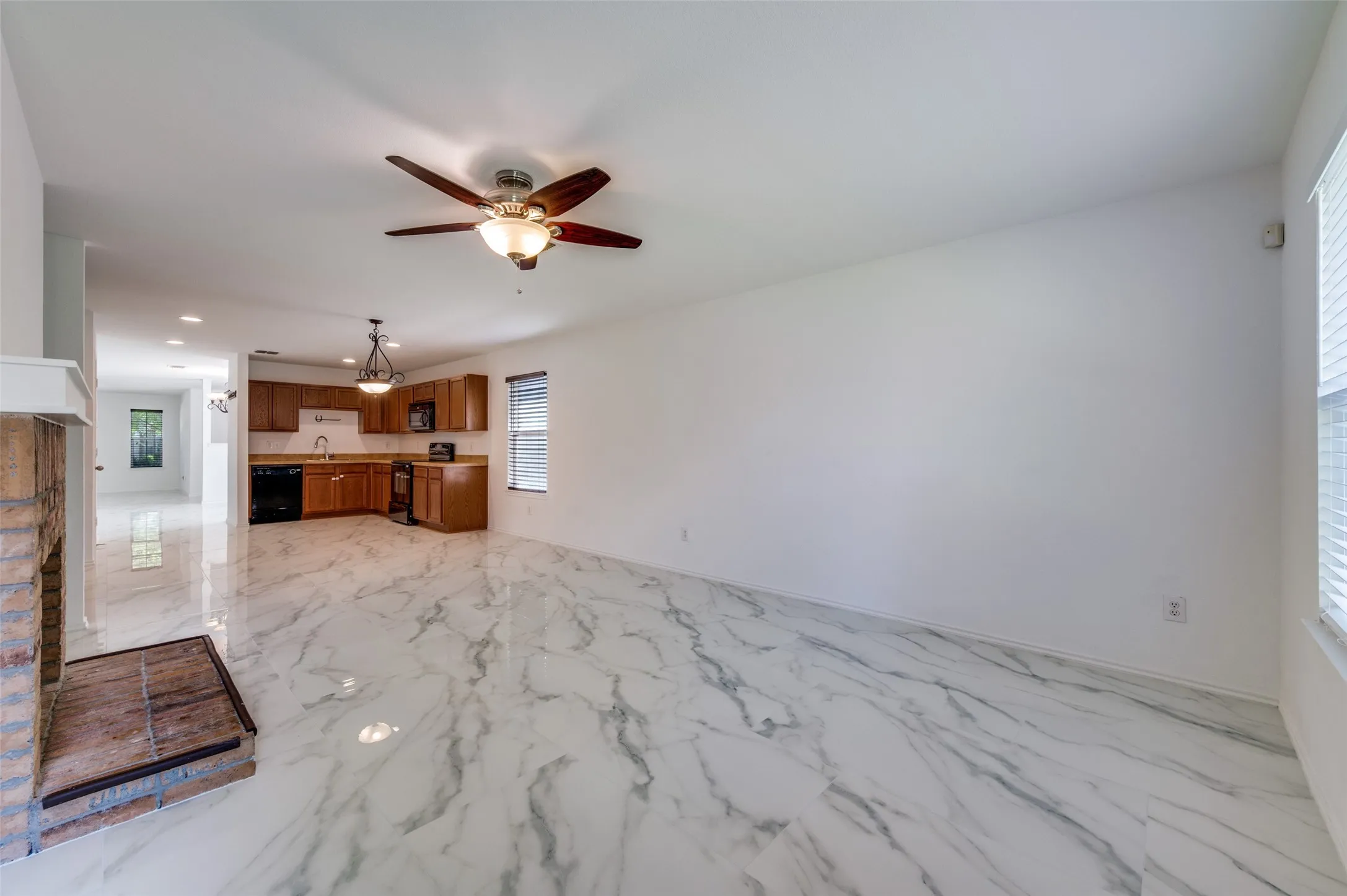 Open-concept living area featuring white marble-look flooring, a brick fireplace with a raised hearth, and a ceiling fan with integrated lighting