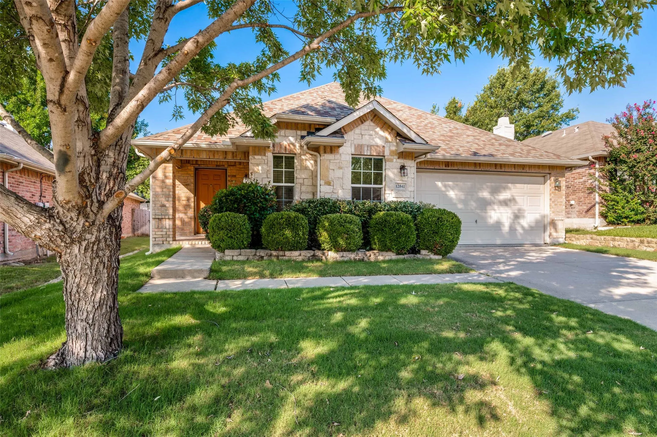 Stone and brick facade home featuring a two-car garage, mature front yard tree, concrete driveway, and established front landscaping