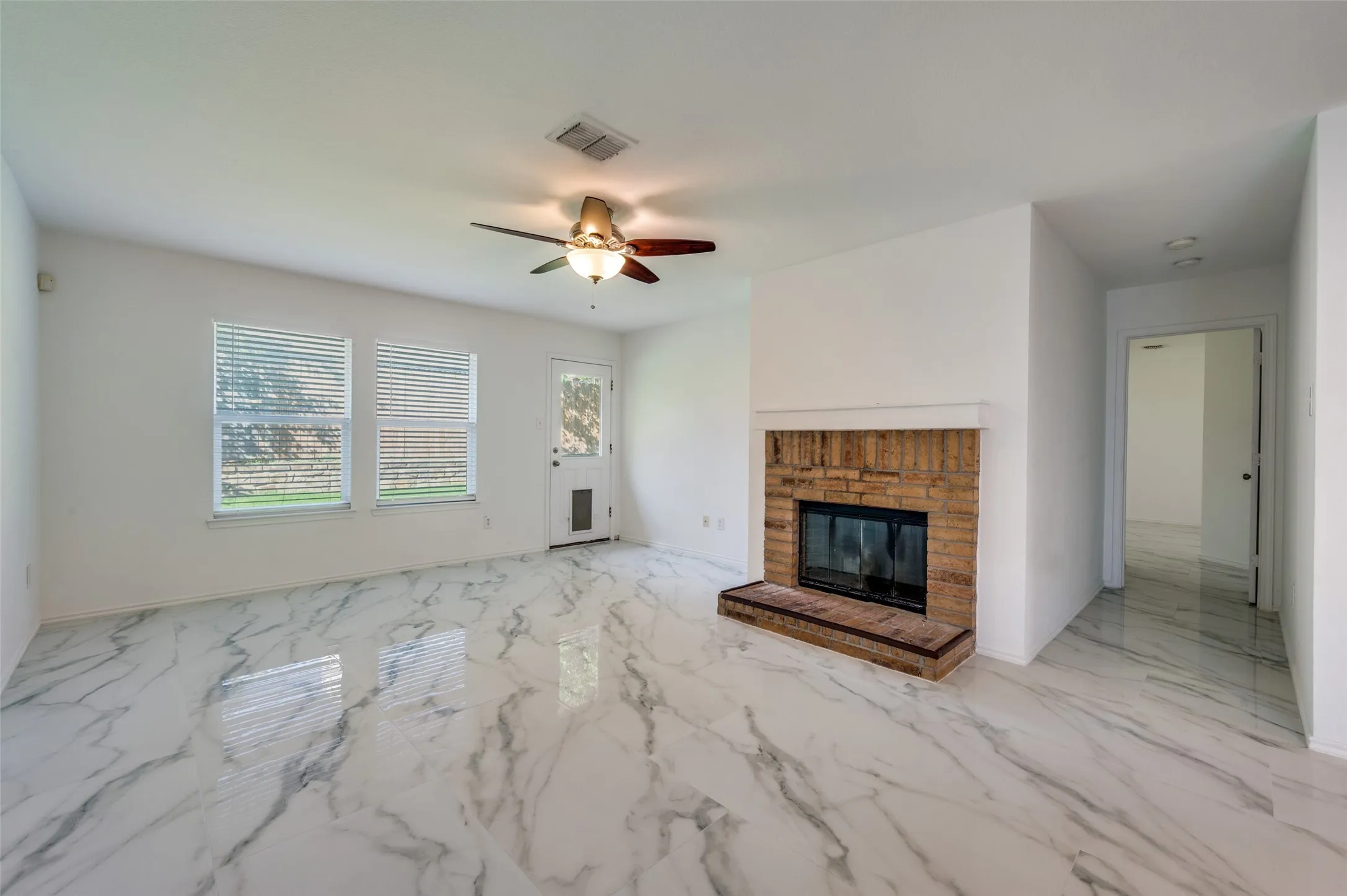 Spacious living area featuring polished marble-look tile flooring, a brick fireplace with hearth, a ceiling fan with integrated lighting, and two windows providing natural light