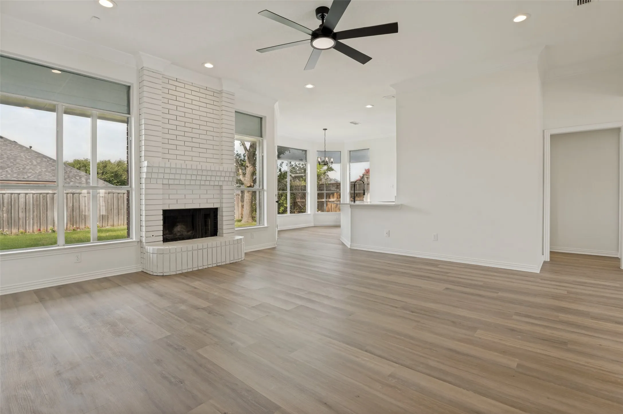 Spacious living area featuring light wood-finish flooring, a white painted brick fireplace, and extensive window coverage