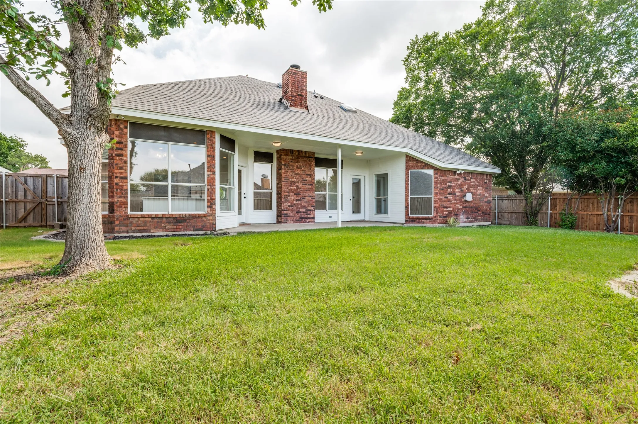 Rear exterior featuring a brick facade, covered patio, multiple large windows, and a fenced yard with mature trees
