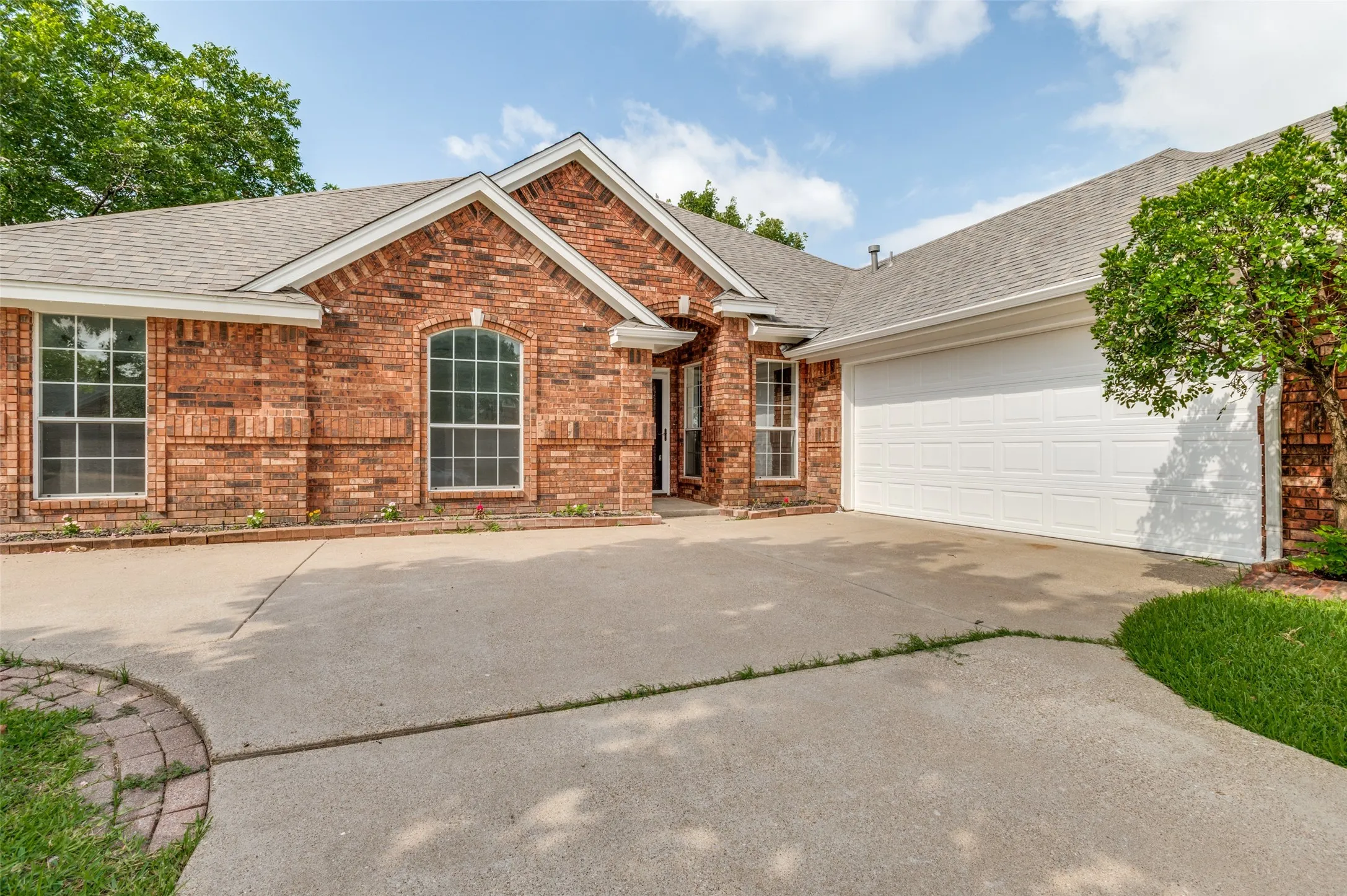 Brick facade residence featuring a prominent arched window, white-trimmed gables, a two-car garage, and a concrete driveway with a brick border