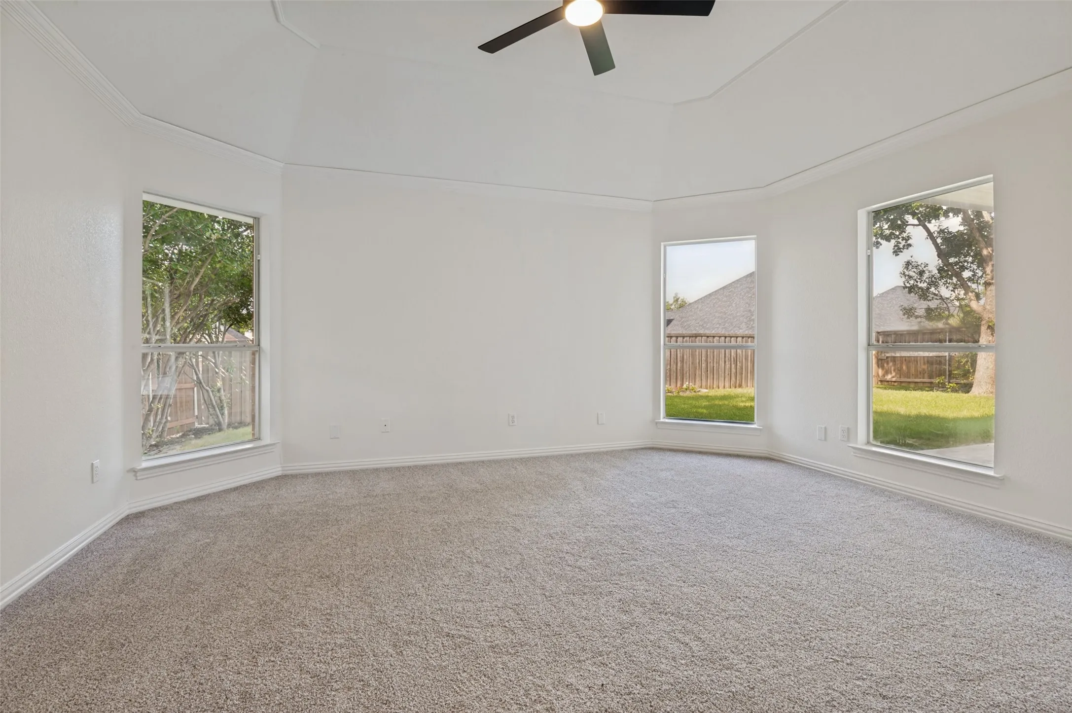 Spacious room featuring light gray carpeting, multiple windows with white trim, a tray ceiling, and a ceiling fan with integrated lighting