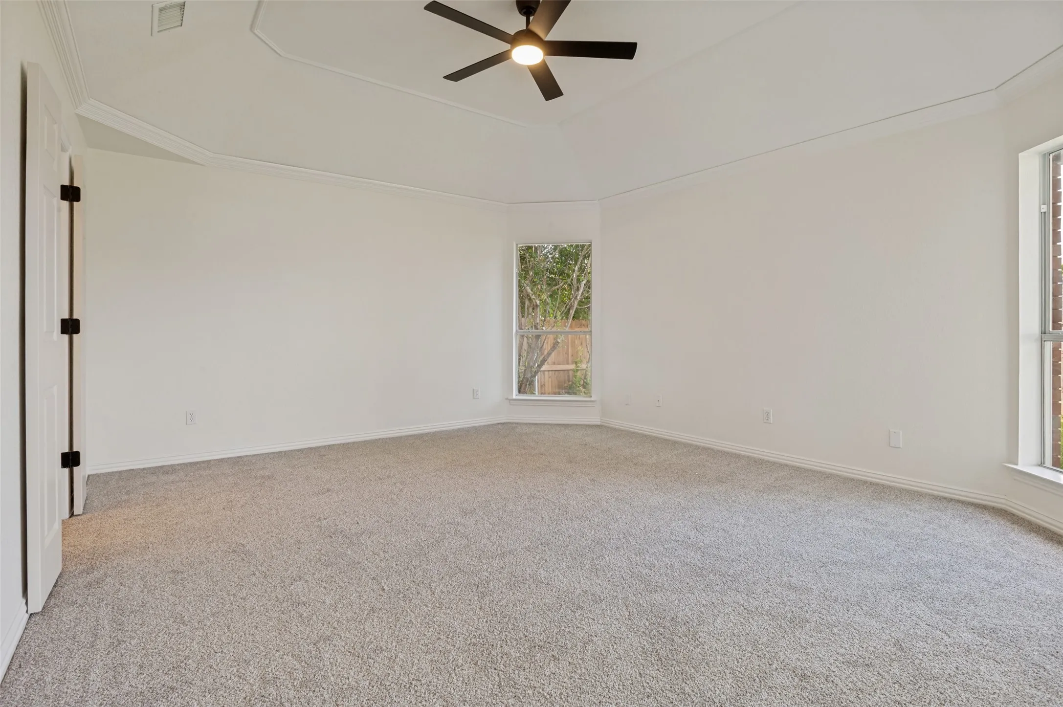 Spacious room featuring light gray carpeting, crown molding, a tray ceiling with a black ceiling fan, and multiple windows providing natural light