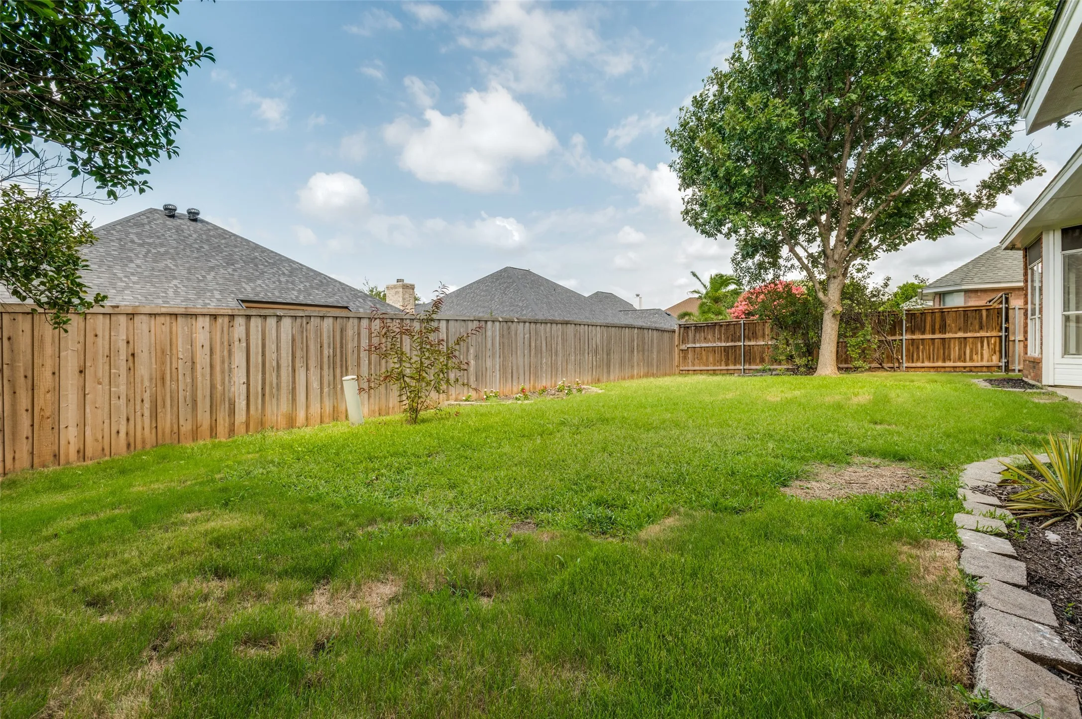 Expansive backyard featuring a large grassy area, a mature tree, perimeter wood fencing, and a stone-edged garden bed
