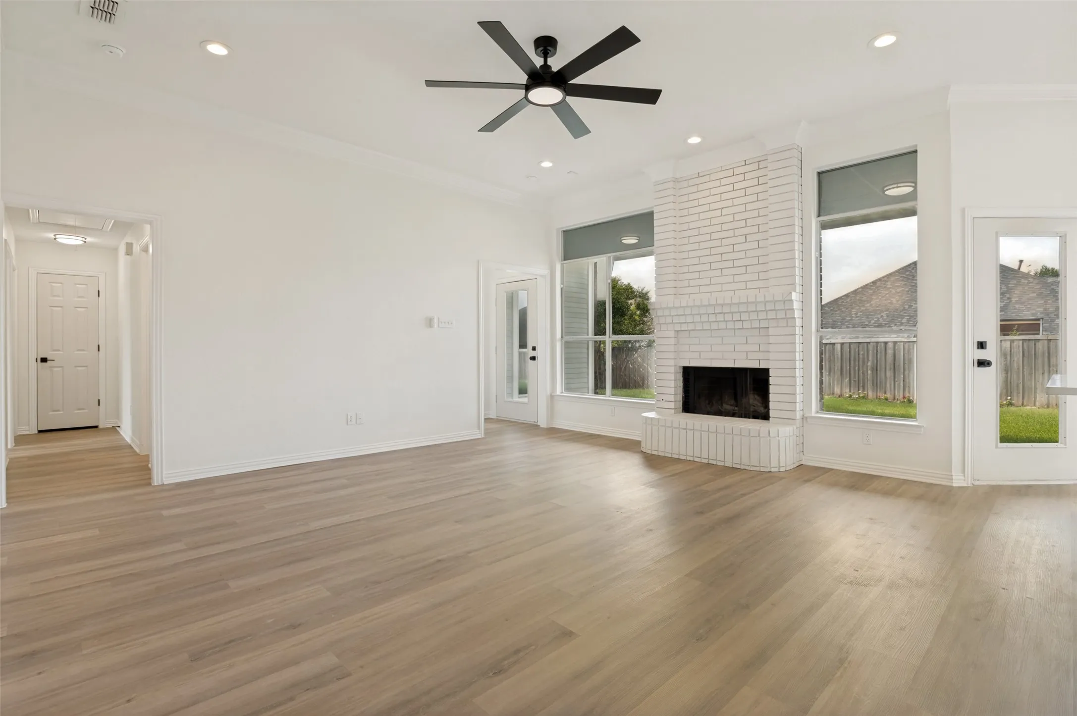 Spacious living area featuring light wood-finish flooring, a white brick fireplace, and ample natural light from multiple windows