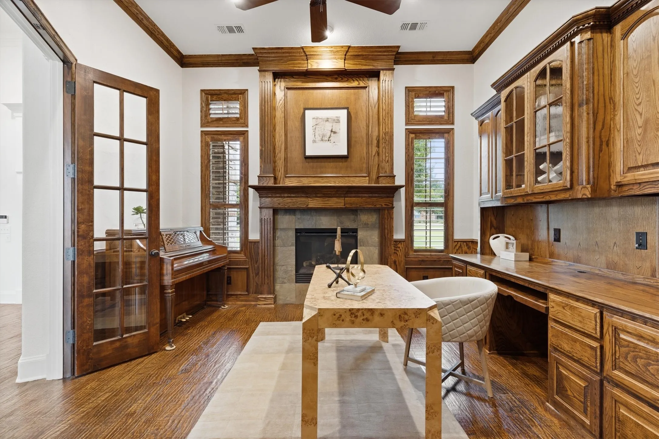 Hardwood flooring throughout, featuring a fireplace with stone surround and wood mantel, built-in desk with extensive cabinetry, wood-framed French doors, and plantation shutters